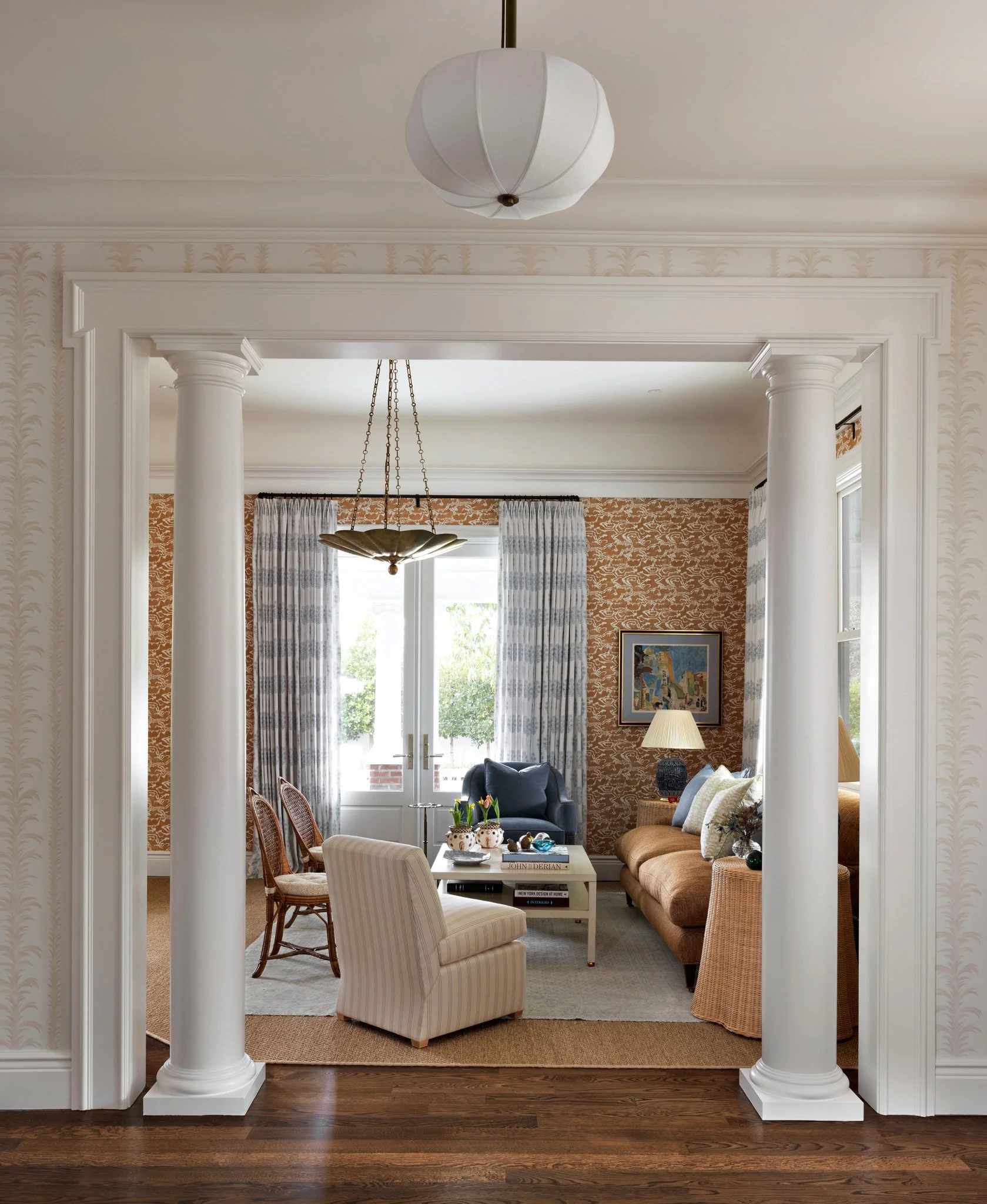 Living room with white columns, a white upholstered armchair, a brown sofa, a coffee table with books and vases, curtains, and a chandelier, viewed through a decorative doorframe.