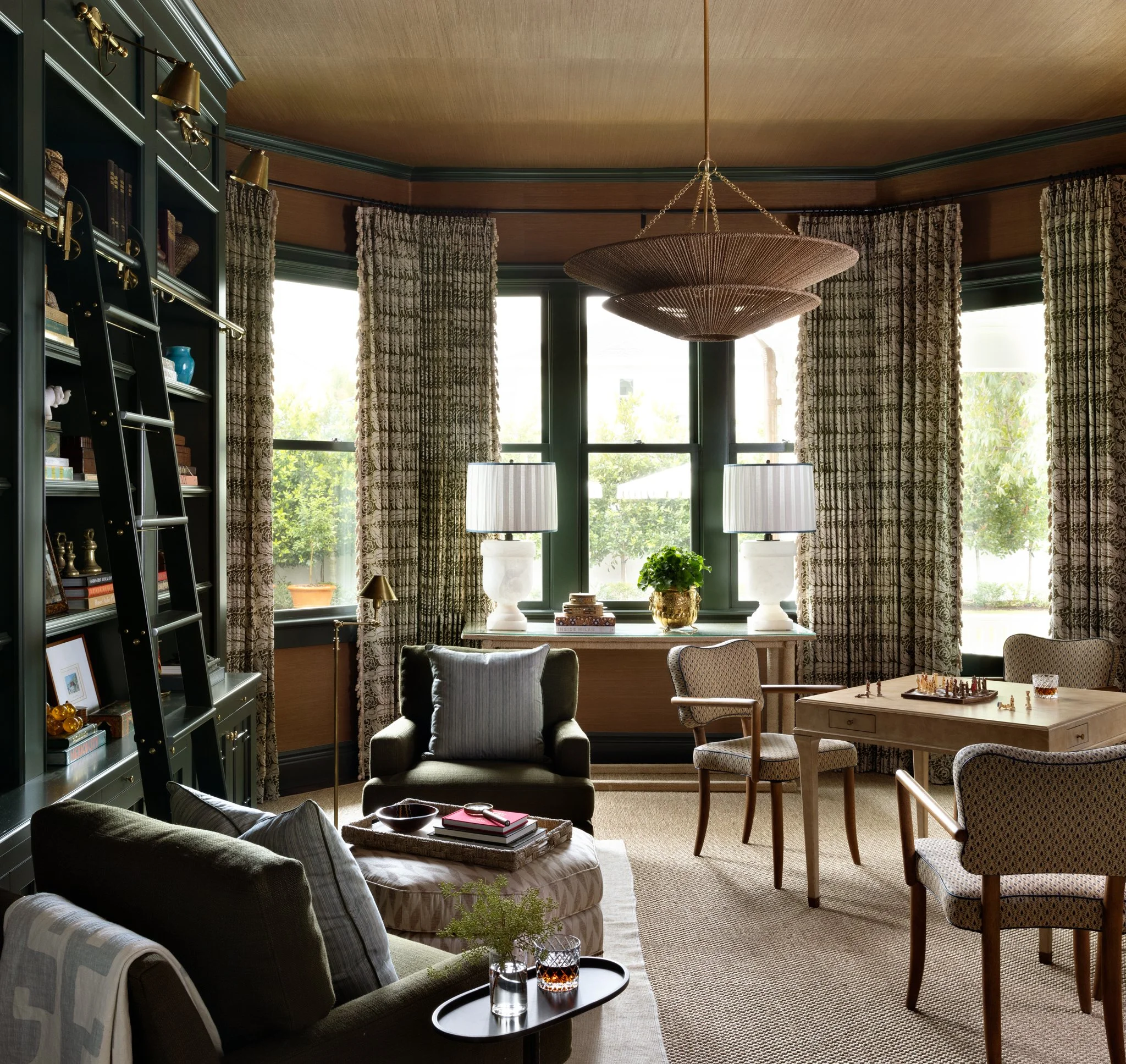 Sunlit living room with large windows, patterned curtains, a dark green bookshelf on the left, a black ladder, upholstered armchair with pillows, a wooden table with two lamps, and a chessboard.