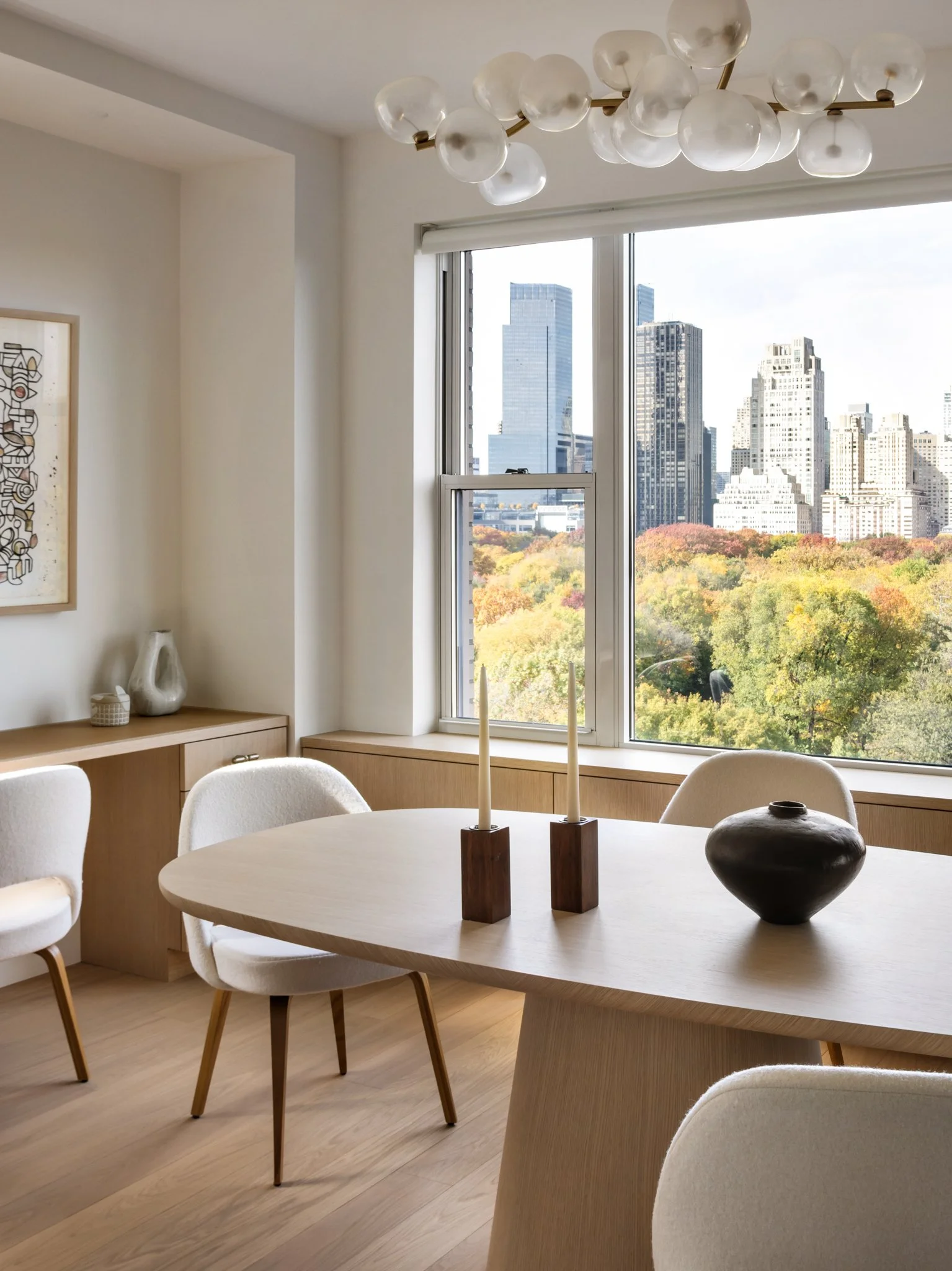 Modern dining room with round table, white upholstered chairs, decorative white vases, wooden candlestick holder, large window with cityscape and trees outside, abstract wall art, brass and glass globe chandelier.