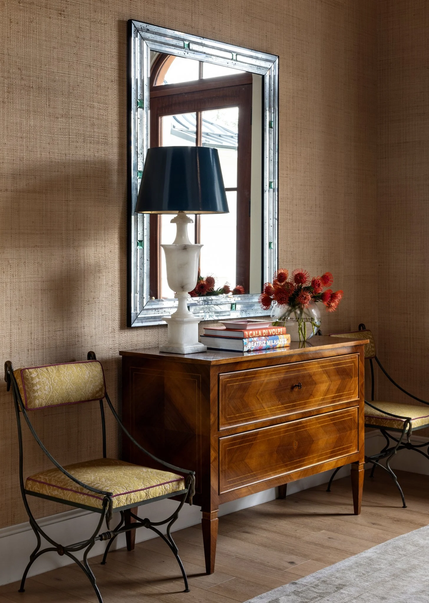 A wooden dresser with a glass vase of orange flowers on top, flanked by two vintage-style chairs with yellow upholstered cushions, a black table lamp, a stack of books, a mirror on the wall, wooden flooring, a beige textured wall, and a window with w