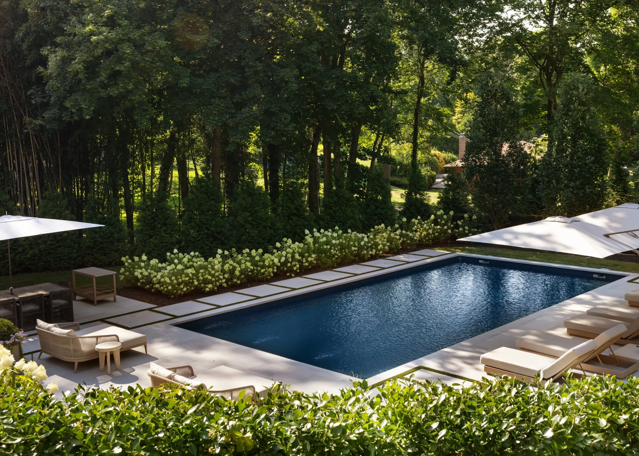 A backyard with a rectangular swimming pool surrounded by white tiles, outdoor lounge chairs, and large umbrellas, with green trees and bushes in the background.