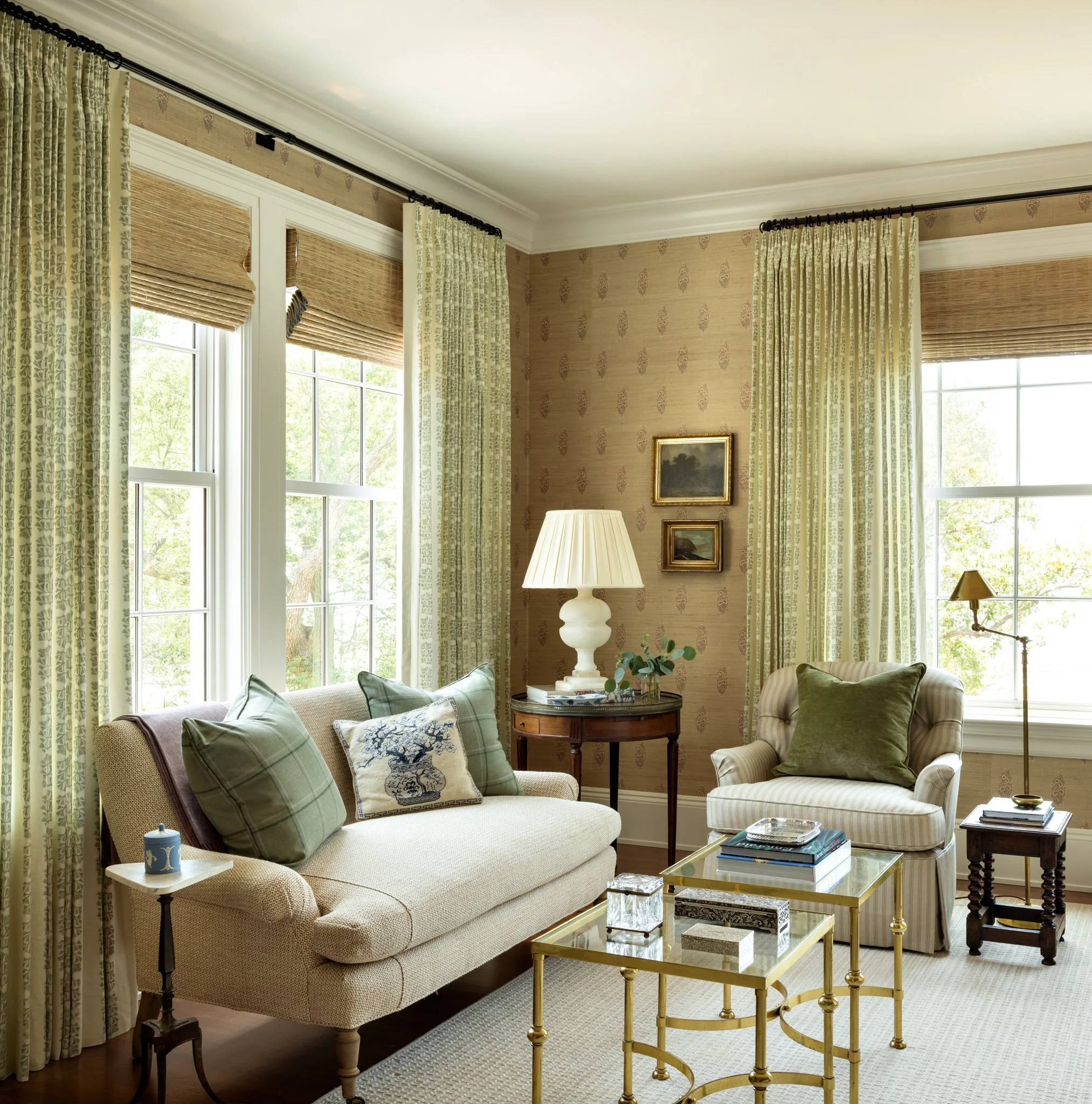Living room with large windows, patterned curtains, beige and green sofas, a glass coffee table with books, a white lamp, framed artwork on the wall, and greenery outside.