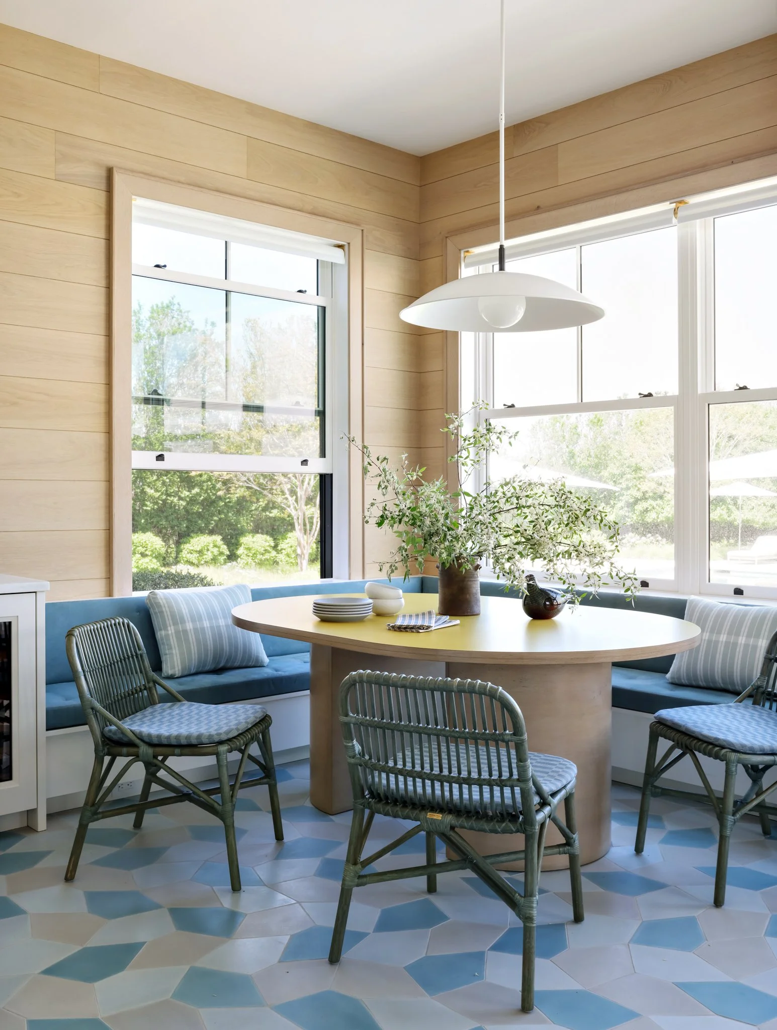 A dining area with a round table, surrounded by chairs with woven pattern, a bench with striped cushions, and a large window revealing a green outdoor landscape, with a vase of greenery and ceramic bowl on the table.