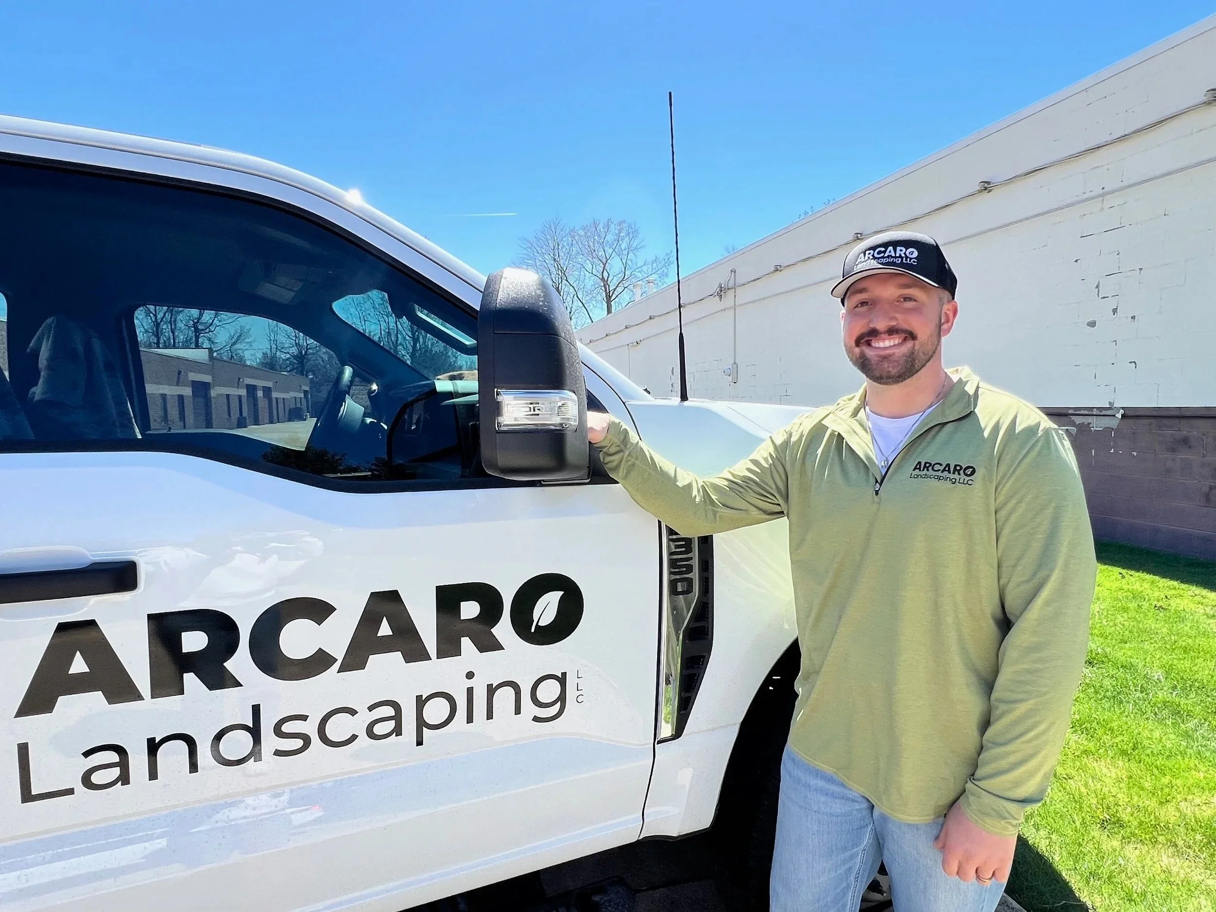 Man standing next to a landscaping company's truck, smiling, wearing a cap and a light green jacket with the company's logo, on a sunny day with blue sky.