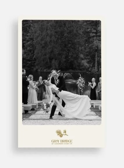 Bride and groom kissing during outdoor wedding ceremony, surrounded by guests clapping. Presented in black and white with wedding venue branding at the bottom.