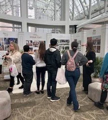 People at an exhibition booth inside a spacious indoor venue with large windows and a glass ceiling, viewing displays and holding bags.