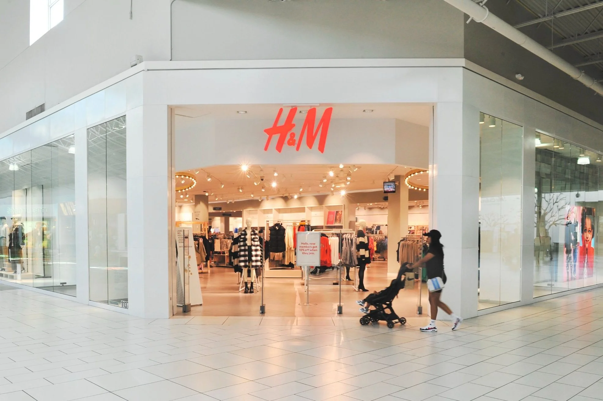 Front entrance of an H&M store in a mall with a woman pushing a stroller.