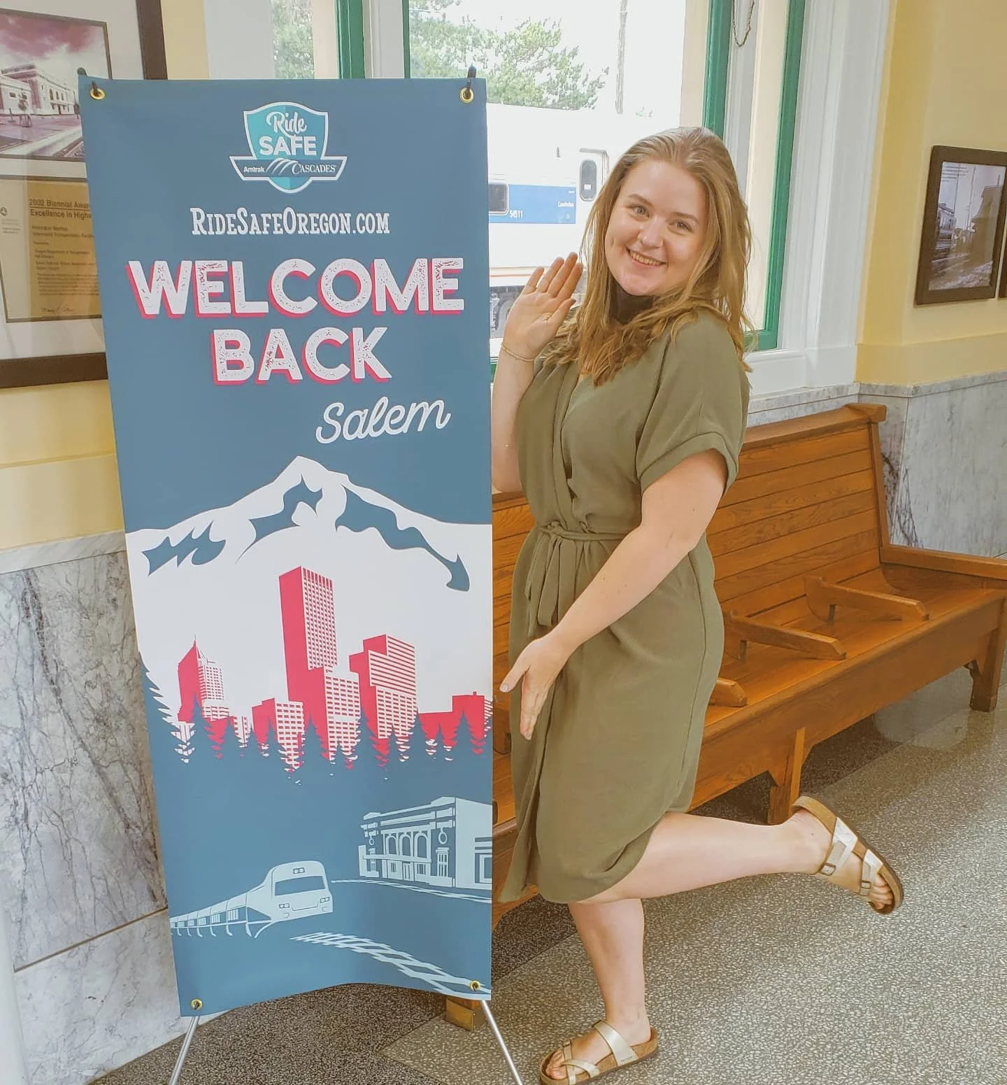 A woman in a green dress stands next to a sign that reads "Welcome Back Salem" with the website address "RideSafeOregon.com." The sign features illustrations of mountains, a city skyline, and a train. The scene appears to be inside a train station with marble walls and wooden benches.