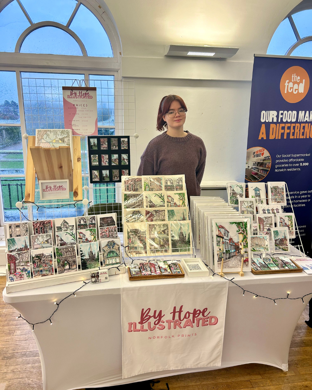 Art display booth with a person standing behind a table full of illustrated prints and artwork. The table has a banner labeled "By Hope Illustrated," and is decorated with string lights. Additional informational banners are visible behind.
