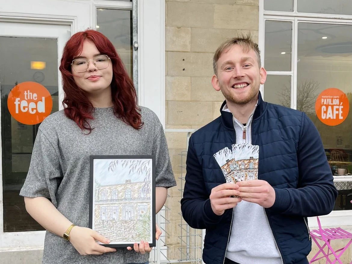 Two people smiling in front of a building with signs reading "the feed" and "waterloo park PAVILION CAFE." One holds a framed drawing, the other holds multiple copies of what appears to be a similar drawing.