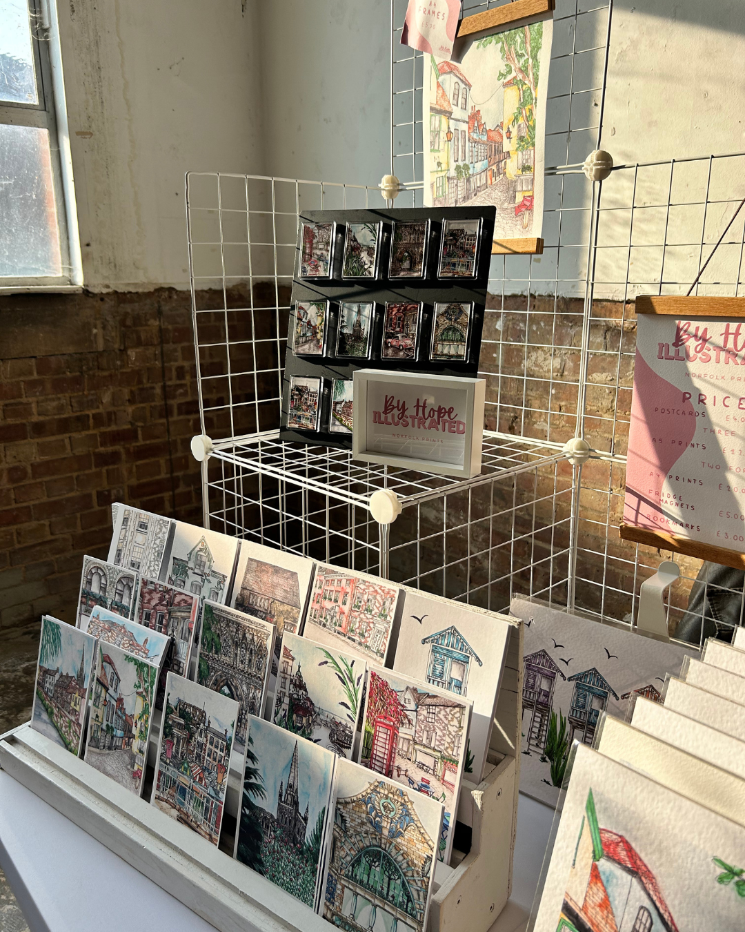 Art display with illustrated postcards and framed art on a white grid stand, featuring colorful architectural designs.