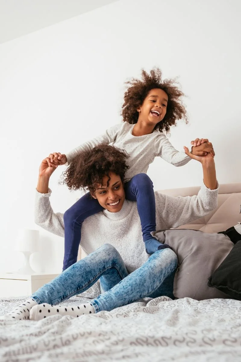 A woman sitting on a bed with two young girls, one sitting on her shoulders and the other holding her hands, all smiling and having fun.