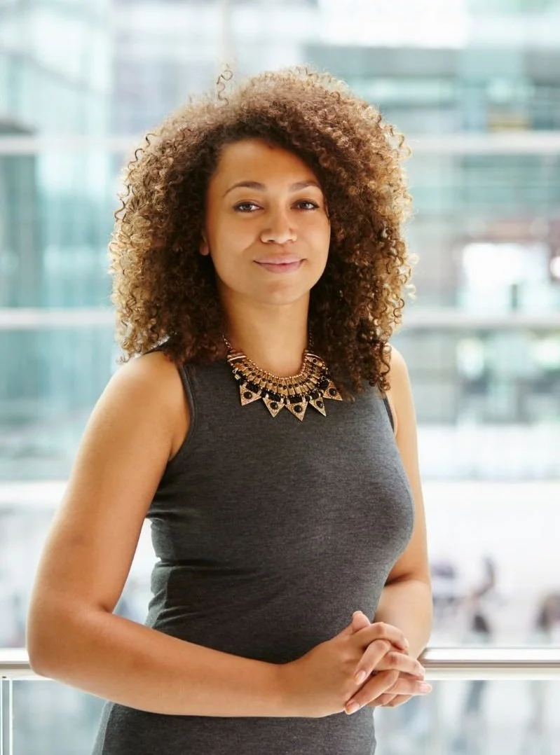 A woman with curly hair wearing a dark gray sleeveless dress and an ornate statement necklace, standing indoors with a glass building exterior visible in the background.