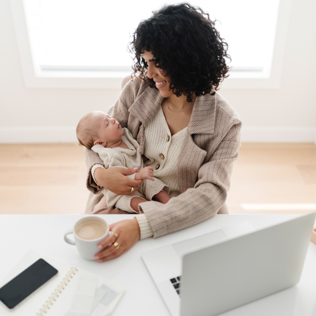 Woman holding a sleeping baby while sitting at a desk with a laptop and coffee in a bright room.