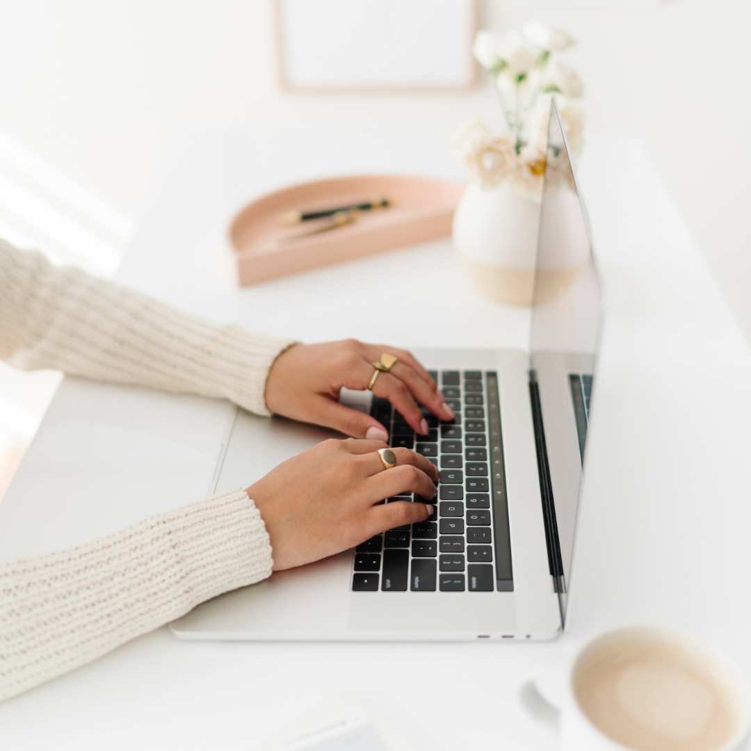 Person wearing a cream-colored sweater typing on a silver laptop at a white desk, with a cup of coffee, a vase with white flowers, and a pink tray in the background.