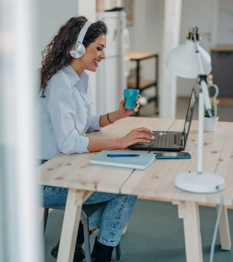 A woman with curly dark hair wearing headphones, smiling while working on a laptop at a wooden desk, holding a blue mug, in a modern office setting.