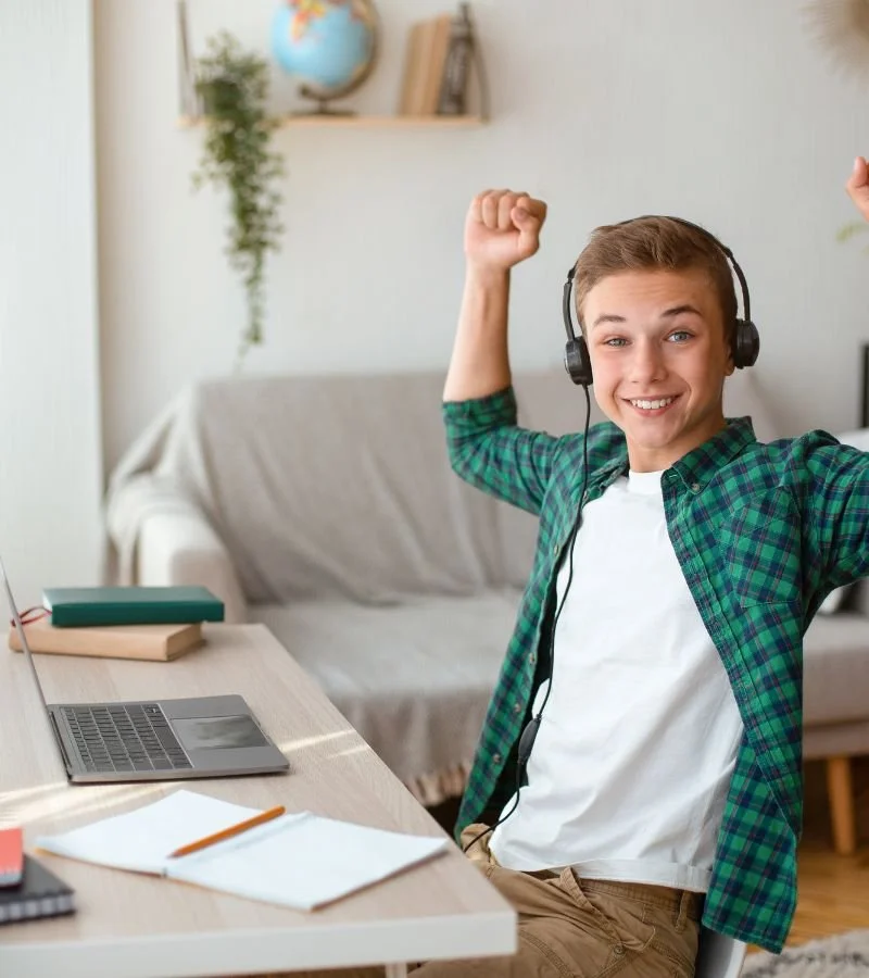 A young man with headphones sitting at a desk, smiling, and flexing his arm muscle in a home setting with books and a computer on the desk.