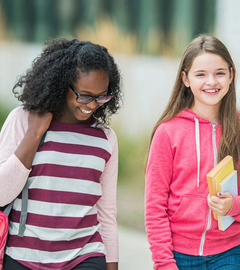 Two smiling girls walking outdoors, one with curly hair and glasses, wearing a striped long sleeve shirt, and the other with straight hair, wearing a pink hoodie, holding books.