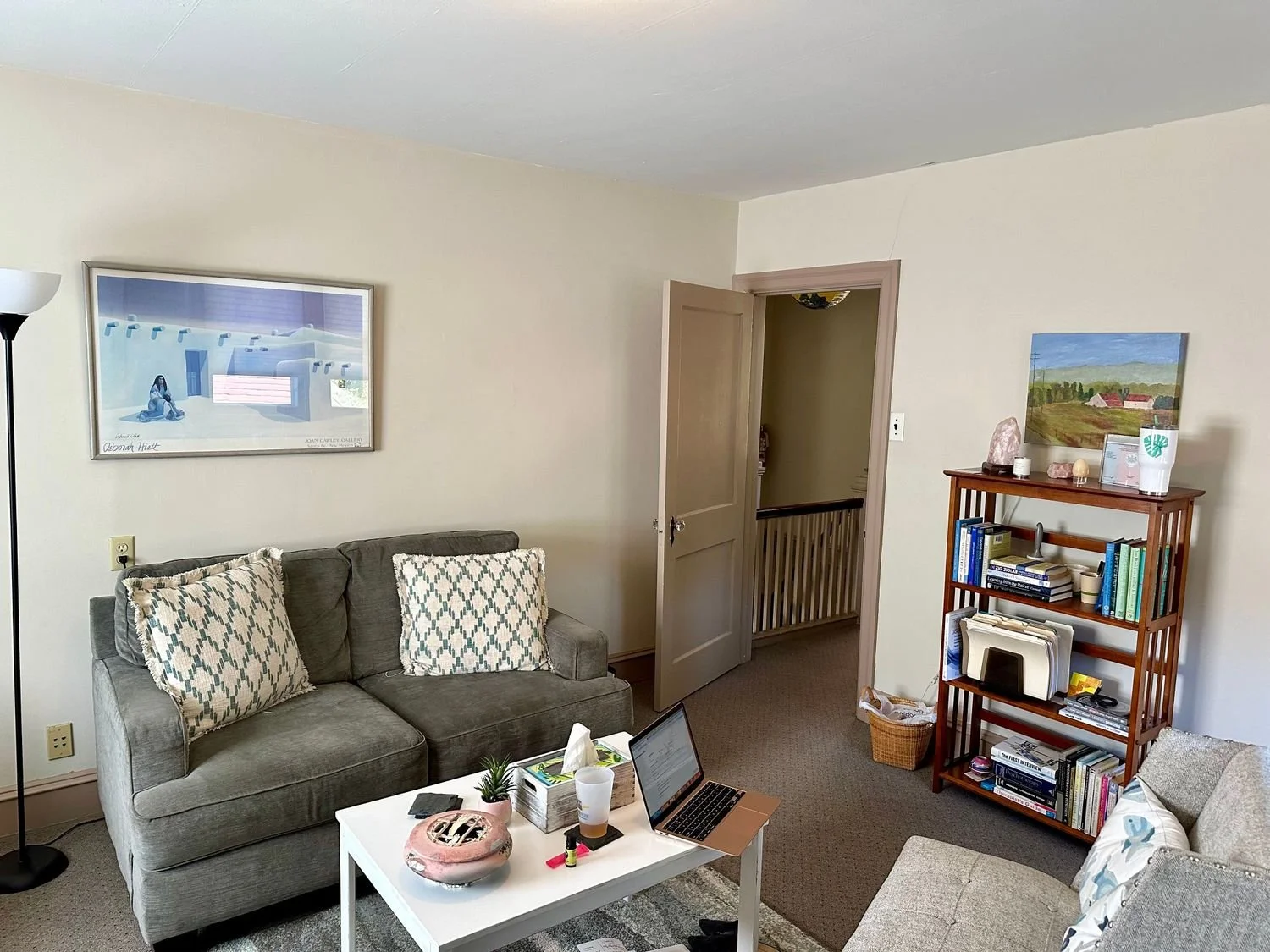 Therapy healing space with a gray sofa, a white coffee table with a small plant, laptop, and various items, a bookshelf with books, a picture hanging on the wall, a door to another room, and a beige armchair with a pillow.