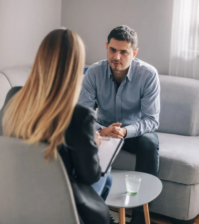 A man sitting on a gray sofa in a therapy or counseling session, talking to a woman who is facing away from the camera, holding a clipboard and a glass of water on a small round table.