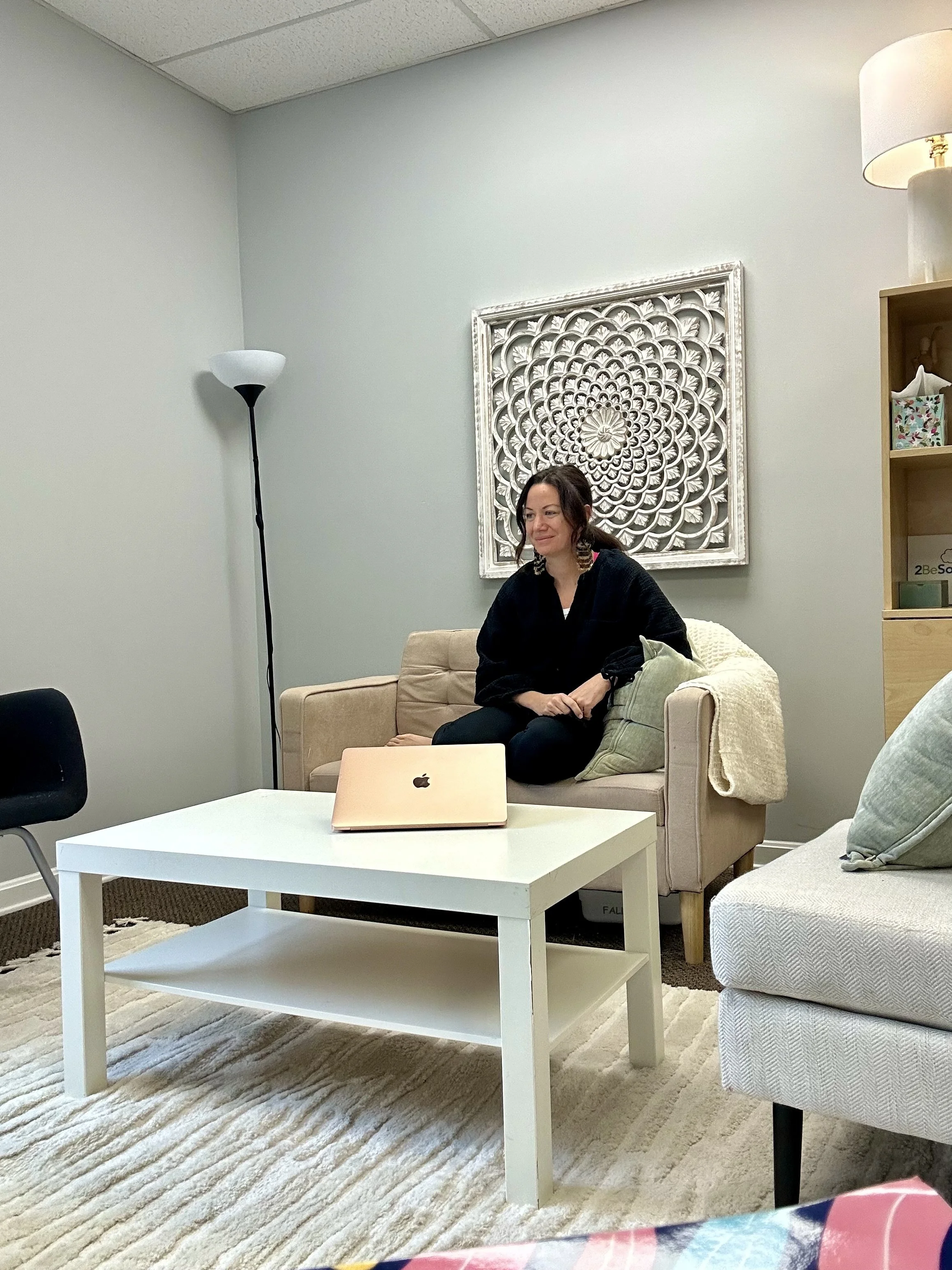 A woman sitting on a beige couch with a cushion, in a room with a white wall, a decorative framed wall art, a tall floor lamp, and a bookshelf. There is a white coffee table with a closed pink laptop on it.