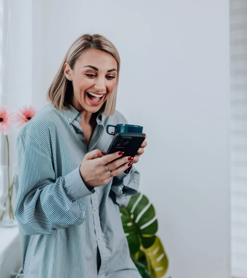 A woman with blonde hair in a striped shirt looks at her phone with a big smile, holding a coffee mug in her left hand, in a bright room with flowers and green plants.