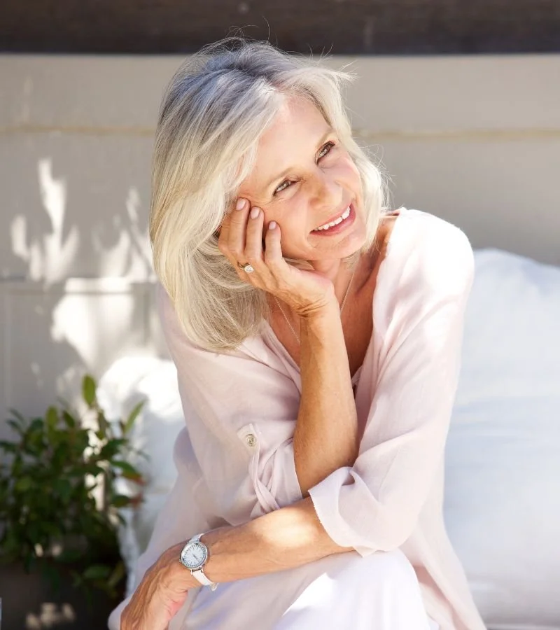 A smiling elderly woman with gray hair sits outdoors on a bright day, resting her head on her hand.