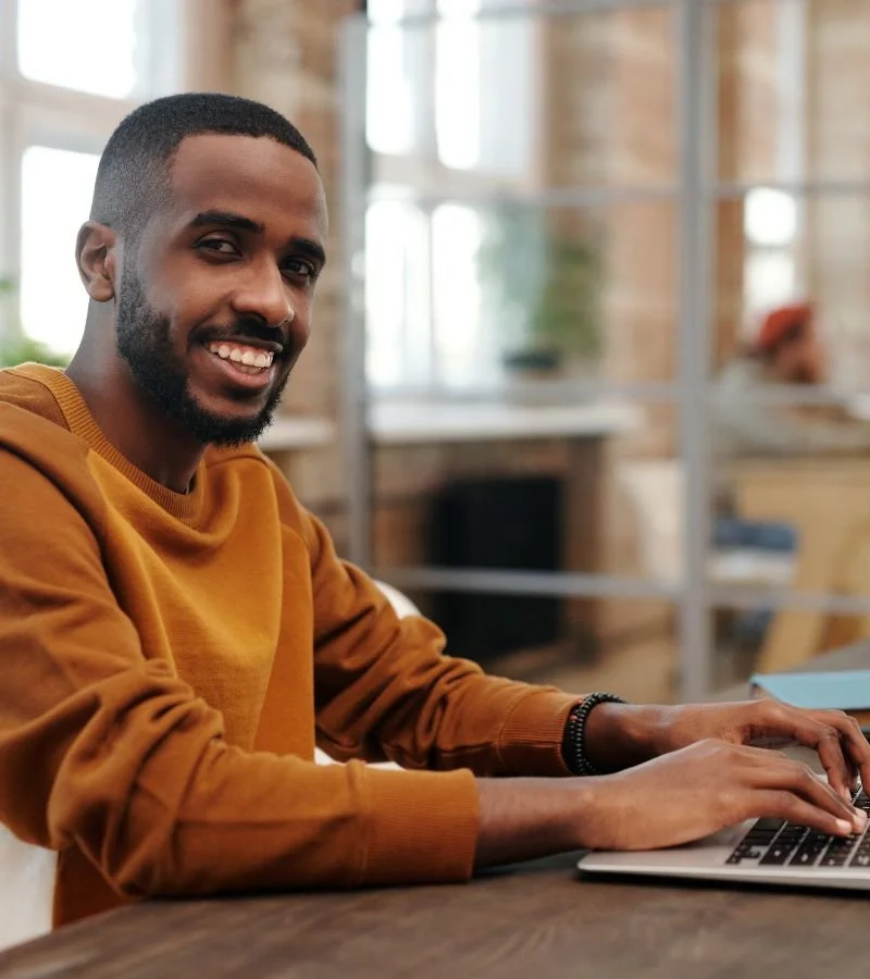A smiling man with a beard and short hair wearing a mustard-colored sweatshirt, sitting at a desk with a laptop in a modern office with large windows and shelves in the background.