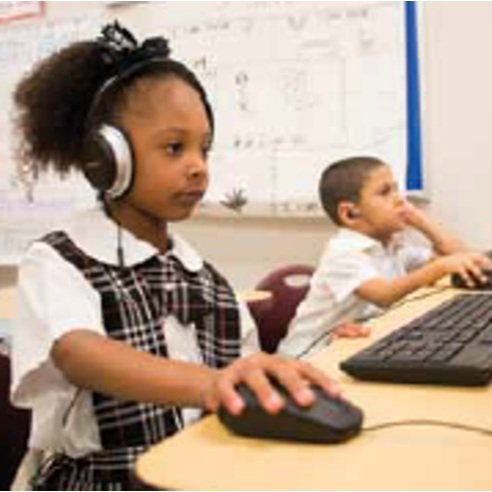 A young girl with headphones working on an UpLift school computer, while a boy in the background listens and interacts with his computer.