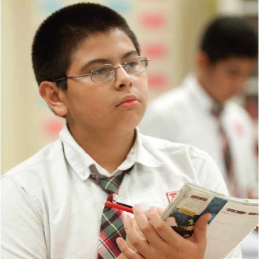 A young boy in a school uniform holding a notebook and pen, looking attentively in an UpLift classroom setting.