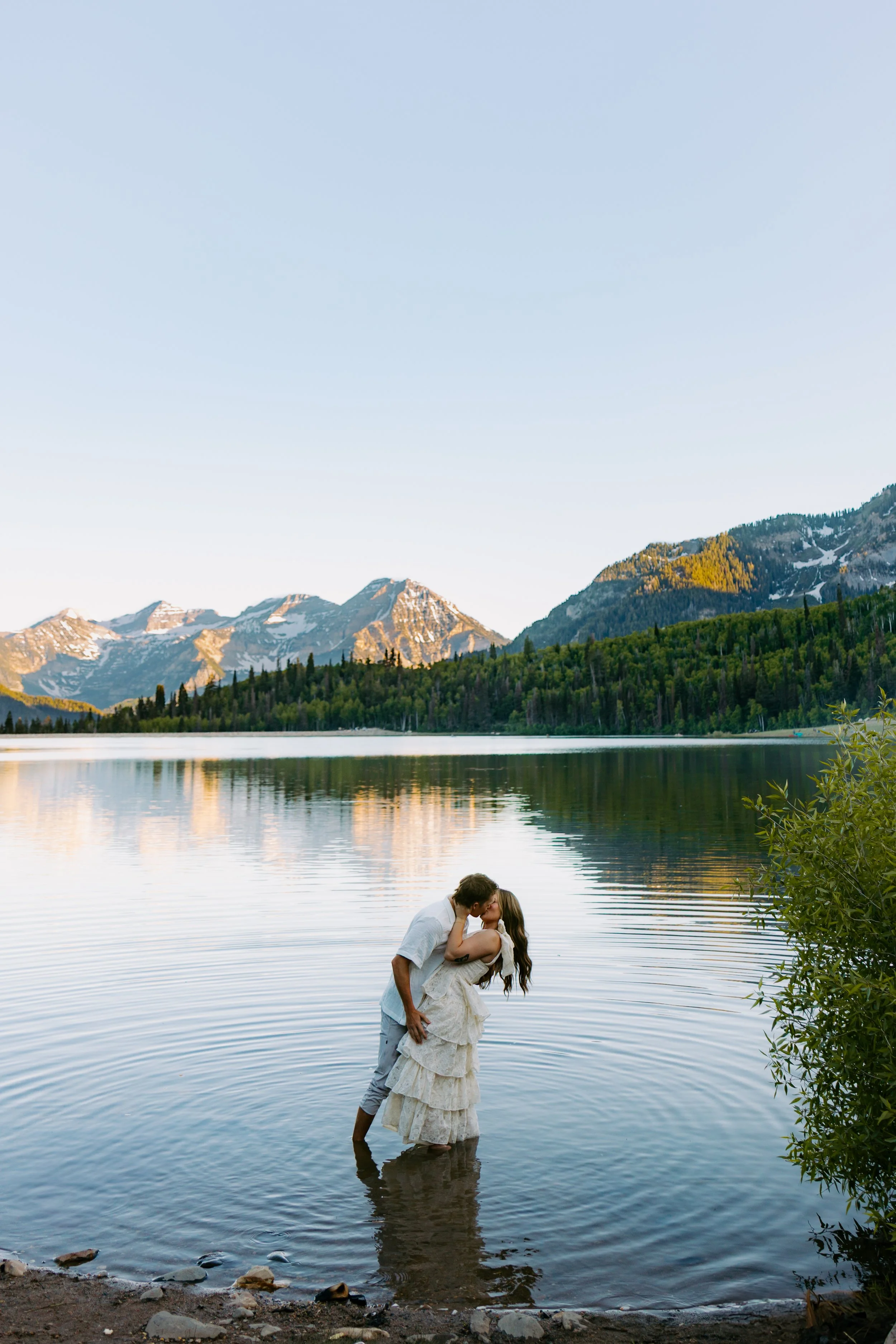 A couple embraces and kisses in a lake with mountains and trees in the background during sunset.
