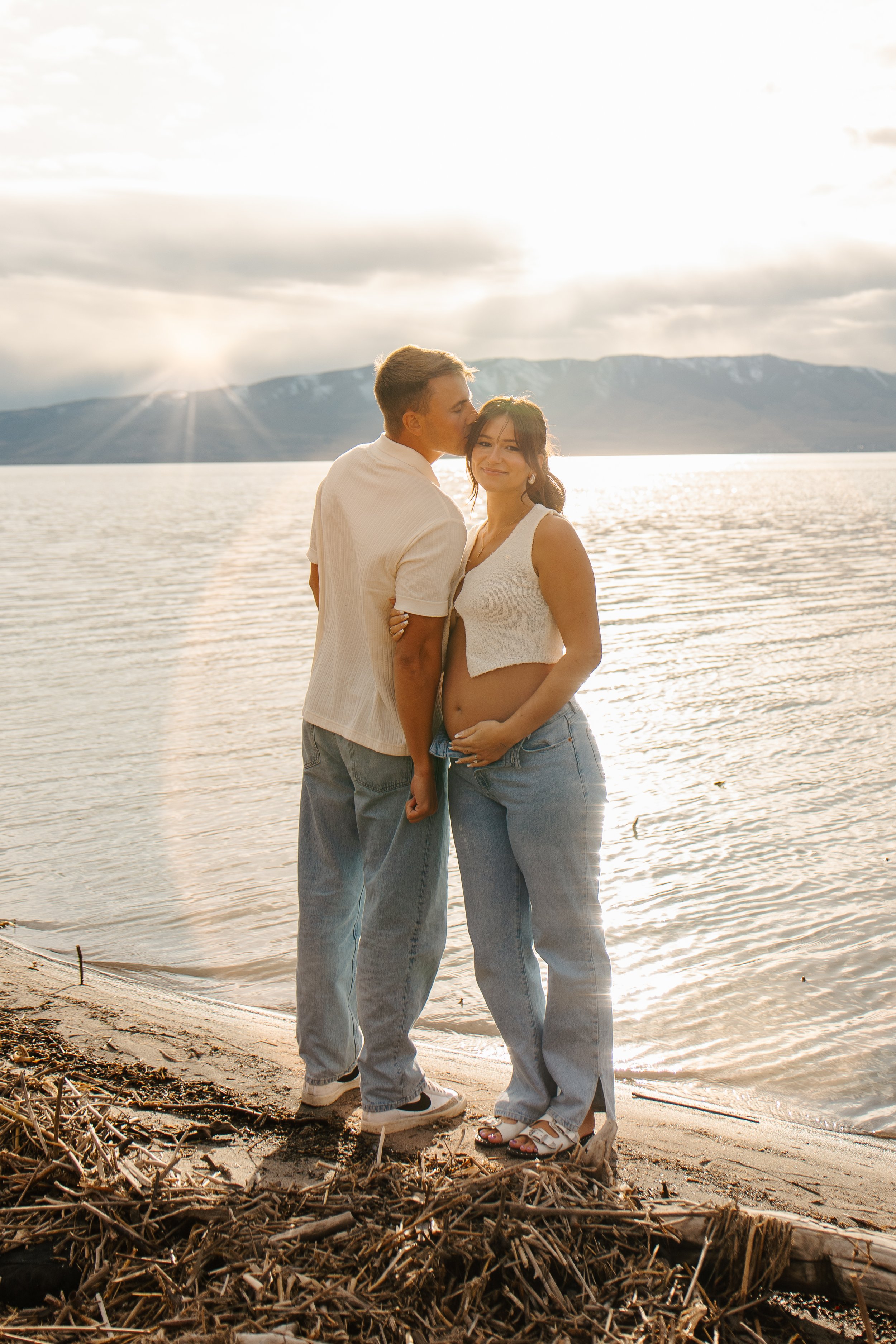 A young couple stands on a beach near a body of water at sunset. The man is kissing the woman on the cheek, and she is holding her pregnant belly, smiling. Mountains are visible in the background.