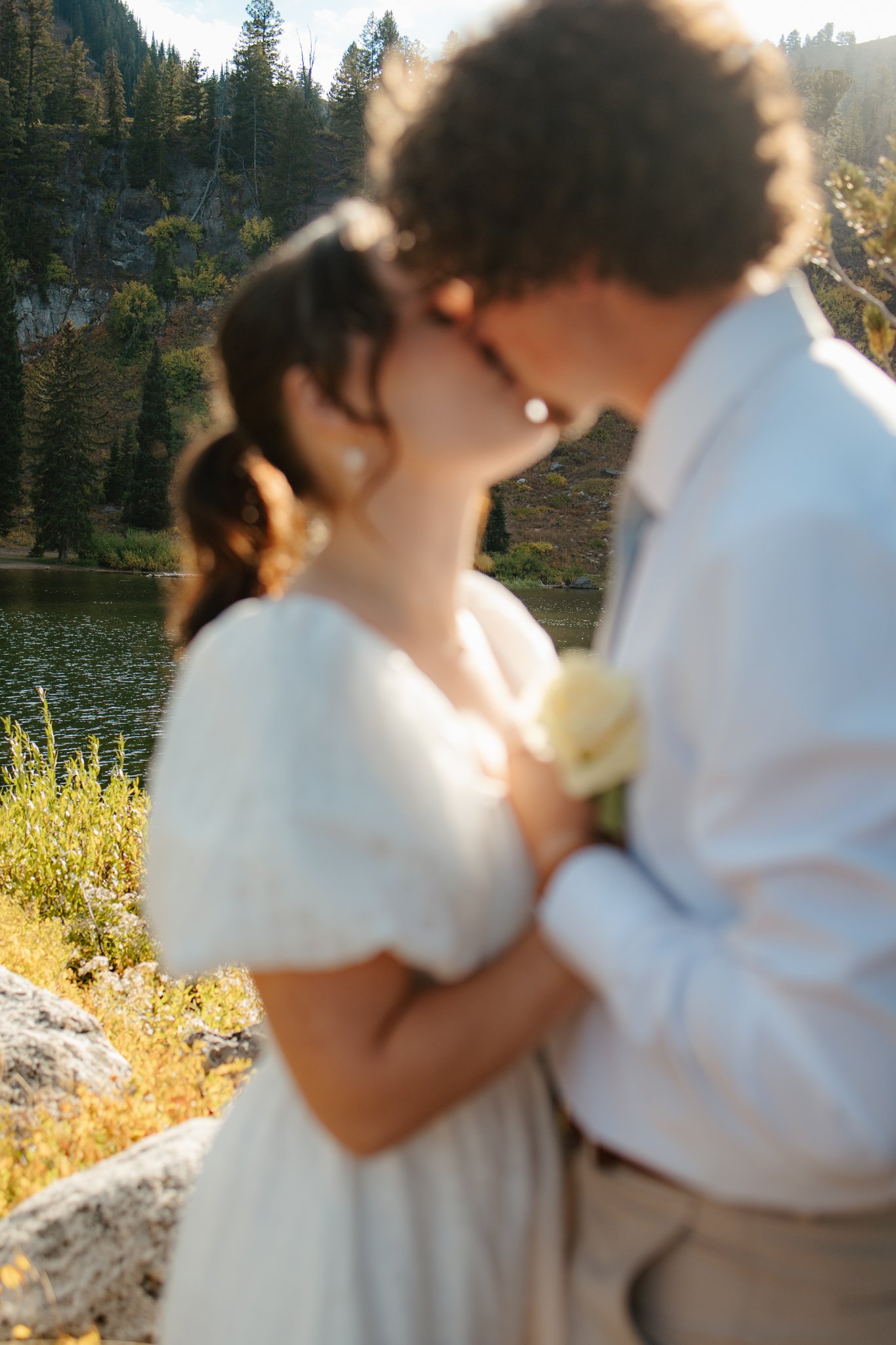 A couple kissing outdoors near a lake and forested hills, with sunlight shining.