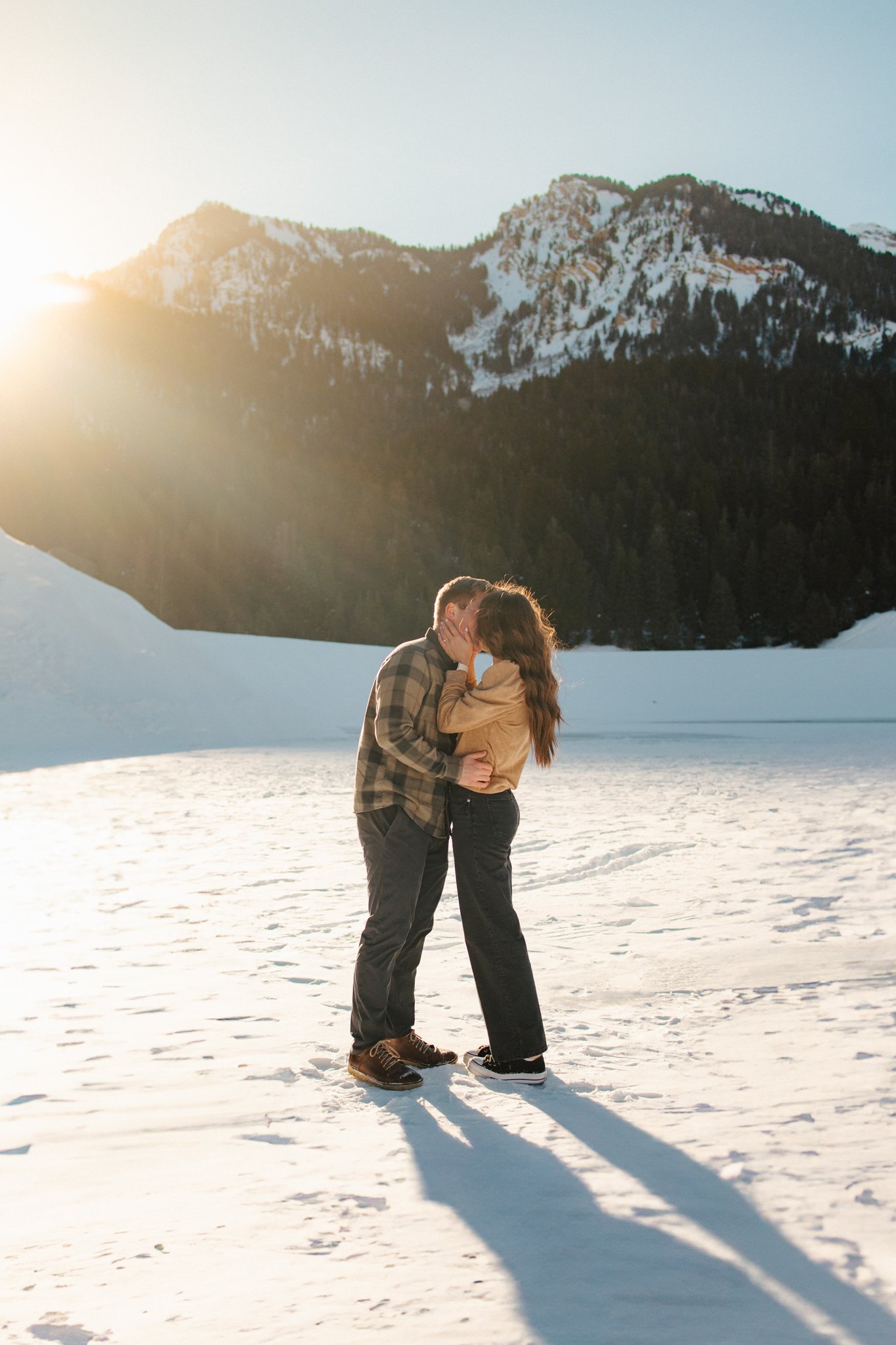 A couple sharing a kiss on a snowy landscape with mountains in the background during sunset.