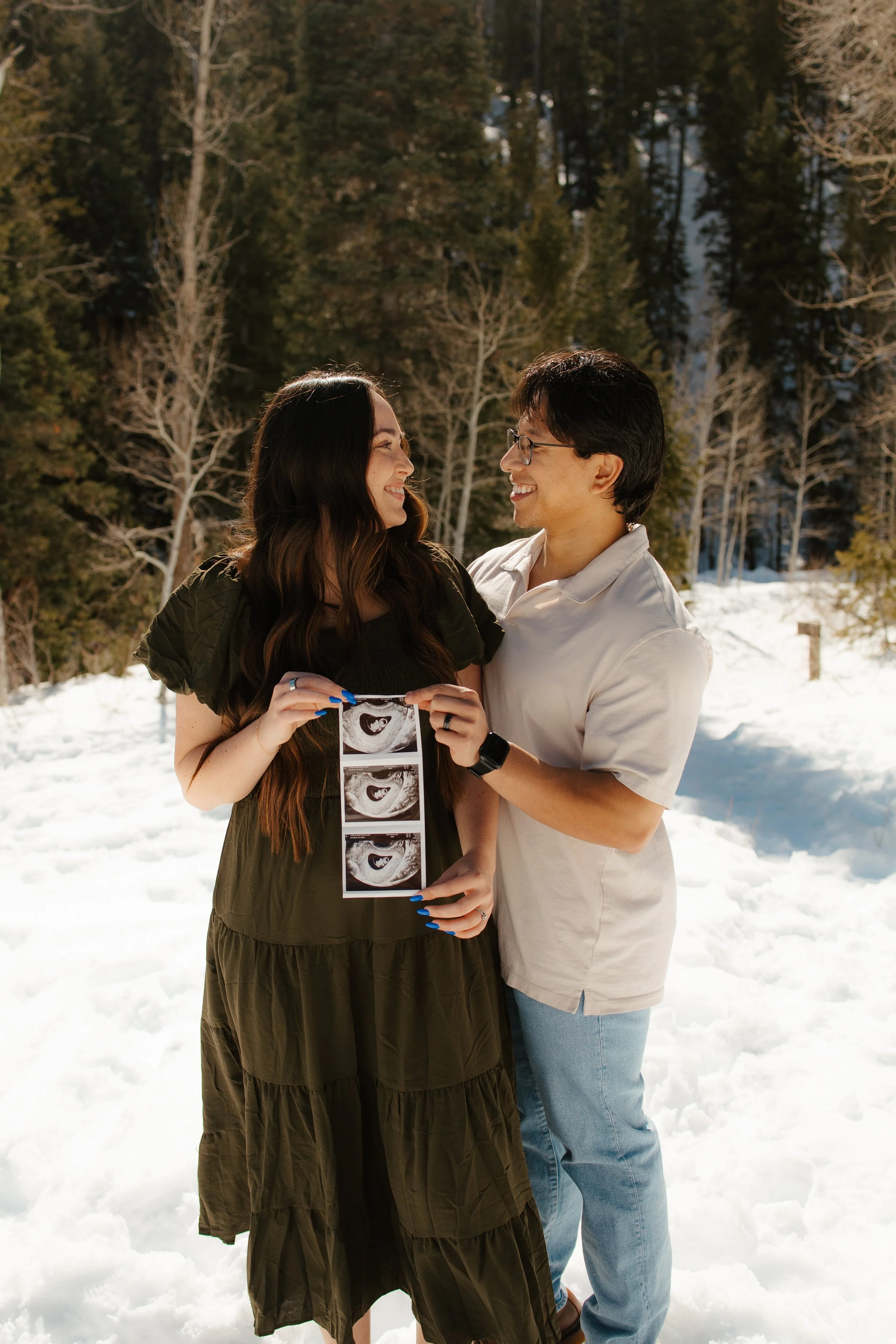 A happy couple standing in a snowy outdoor setting, holding ultrasound images of a baby during daytime.