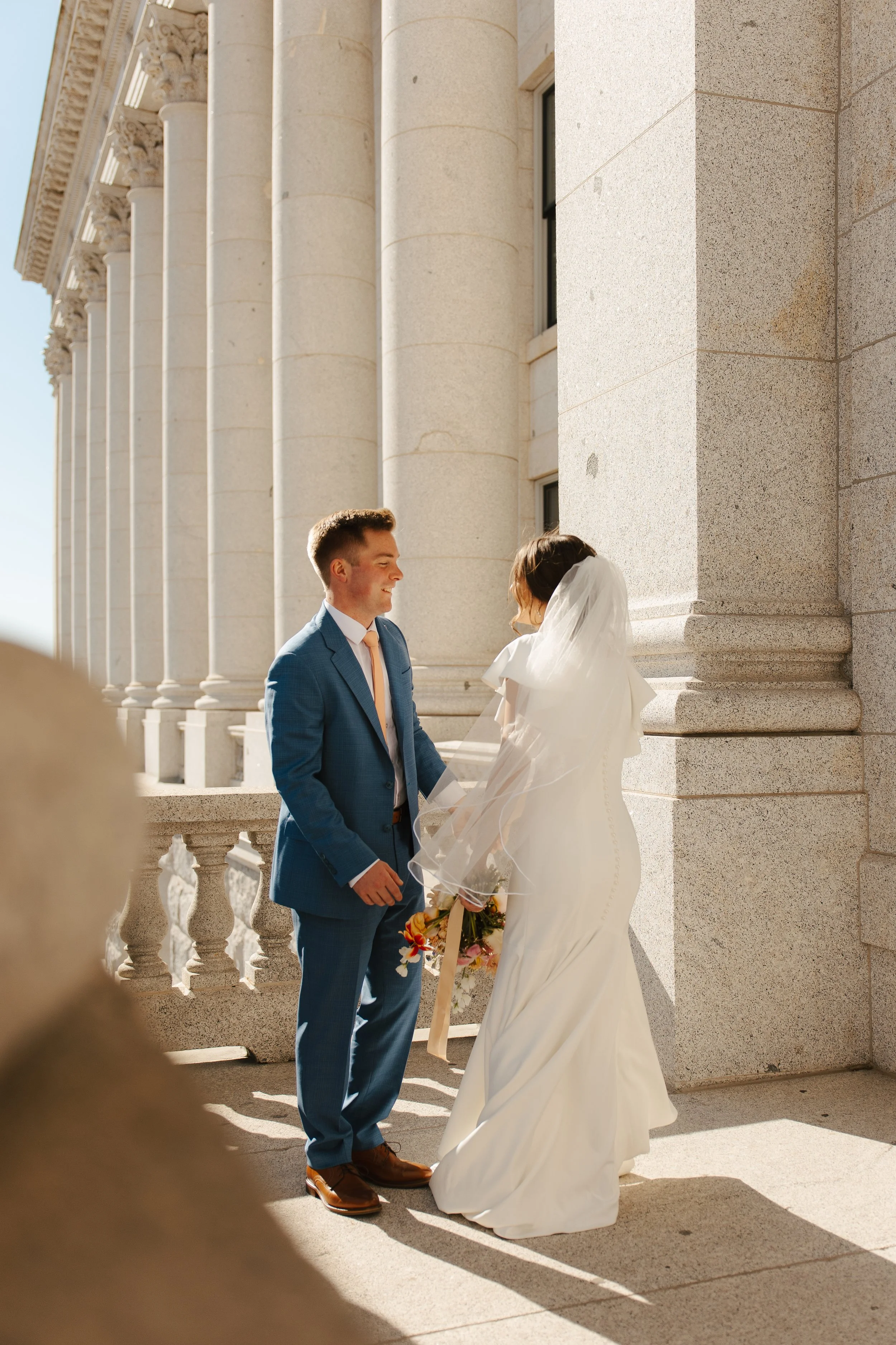 A bride and groom standing outdoors, exchanging vows or a kiss, in front of a grand stone building with large columns, on a sunny day.