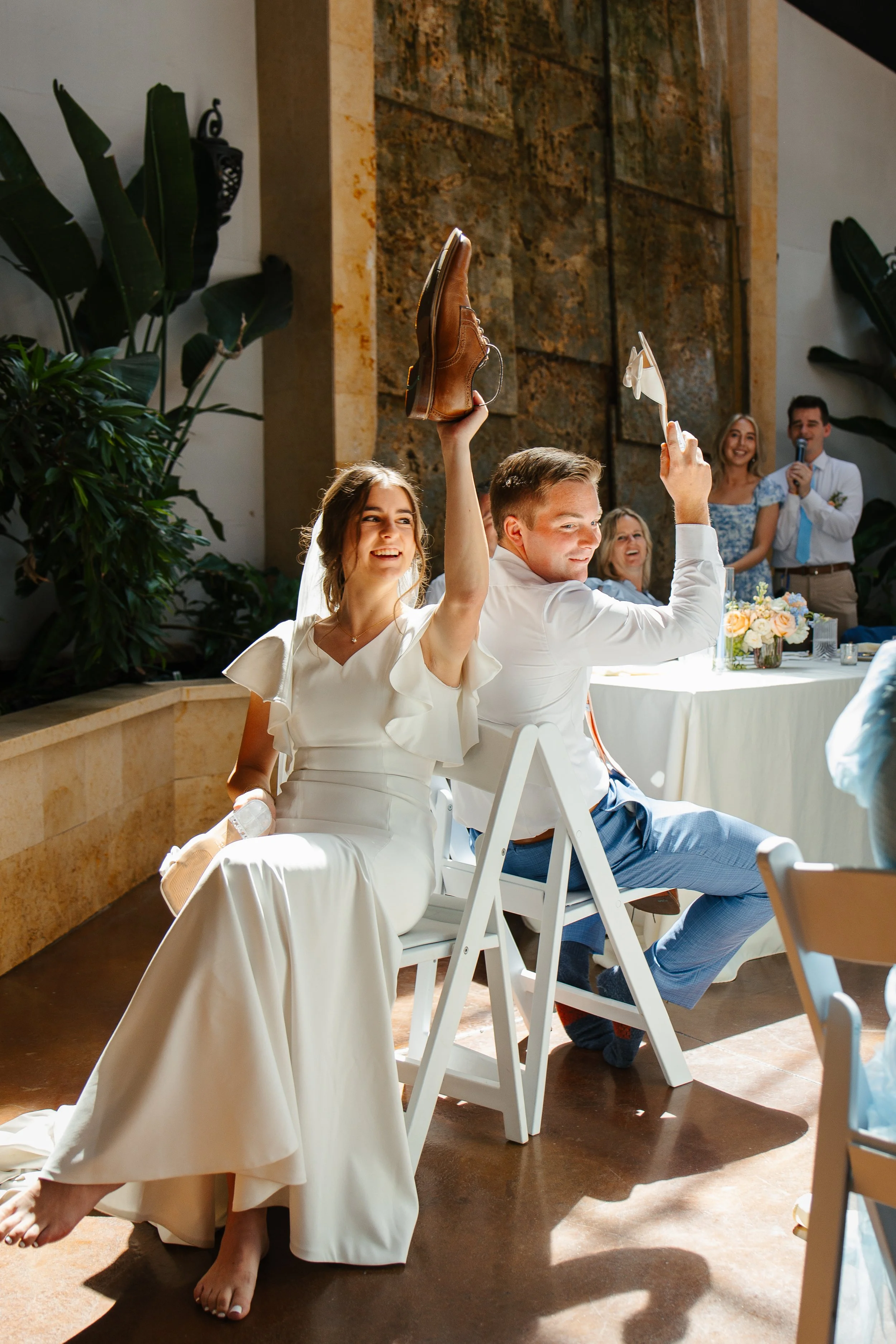 A bride and groom sitting back to back at a wedding reception, raising shoes and a sandal in celebration, with guests smiling behind them.