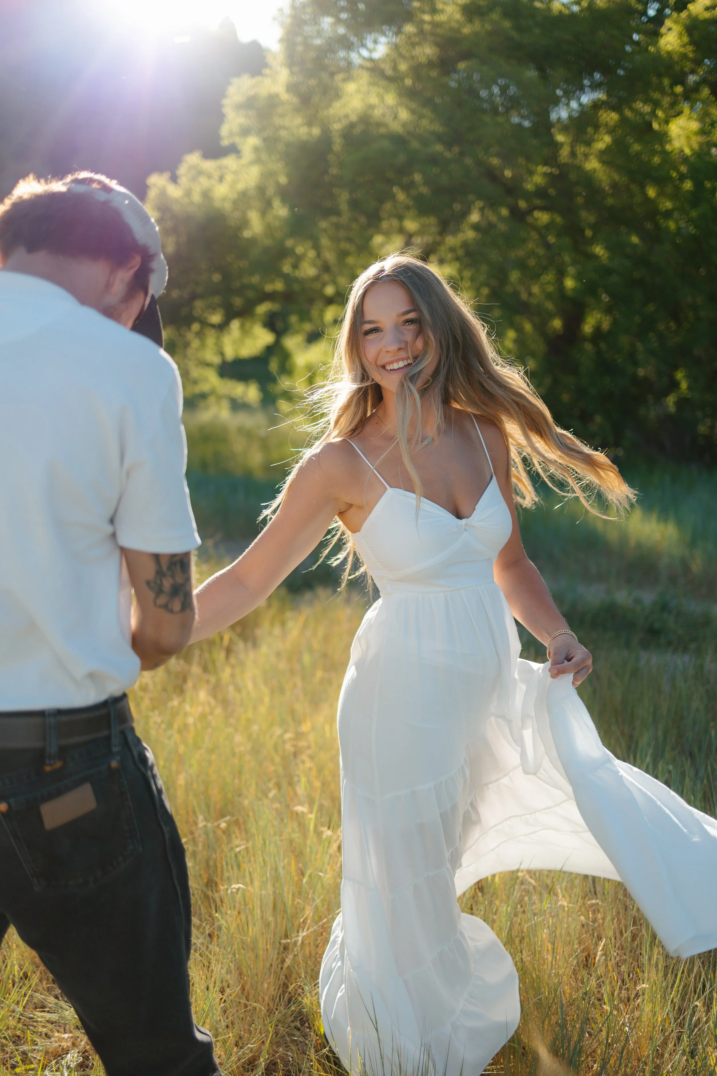 A young woman in a white dress smiling and playing in a sunlit grassy field, holding the hand of a man whose back is to the camera, with green trees and sunlight in the background.