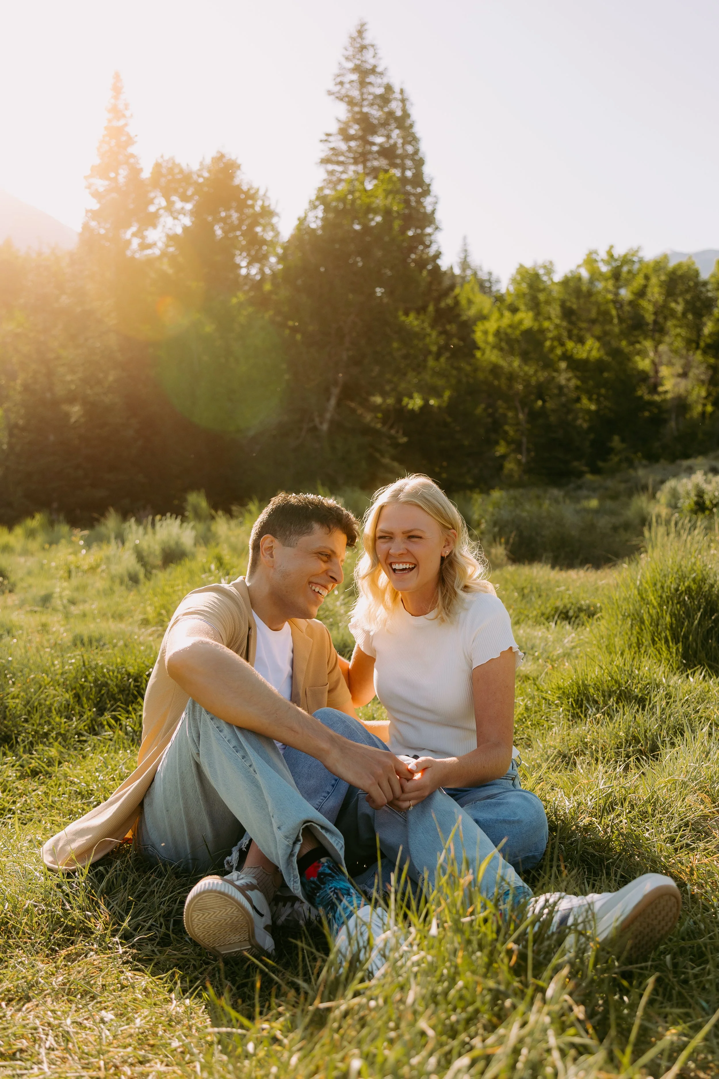 A young couple sitting on grass in a field, laughing and enjoying each other's company on a sunny day with trees in the background.