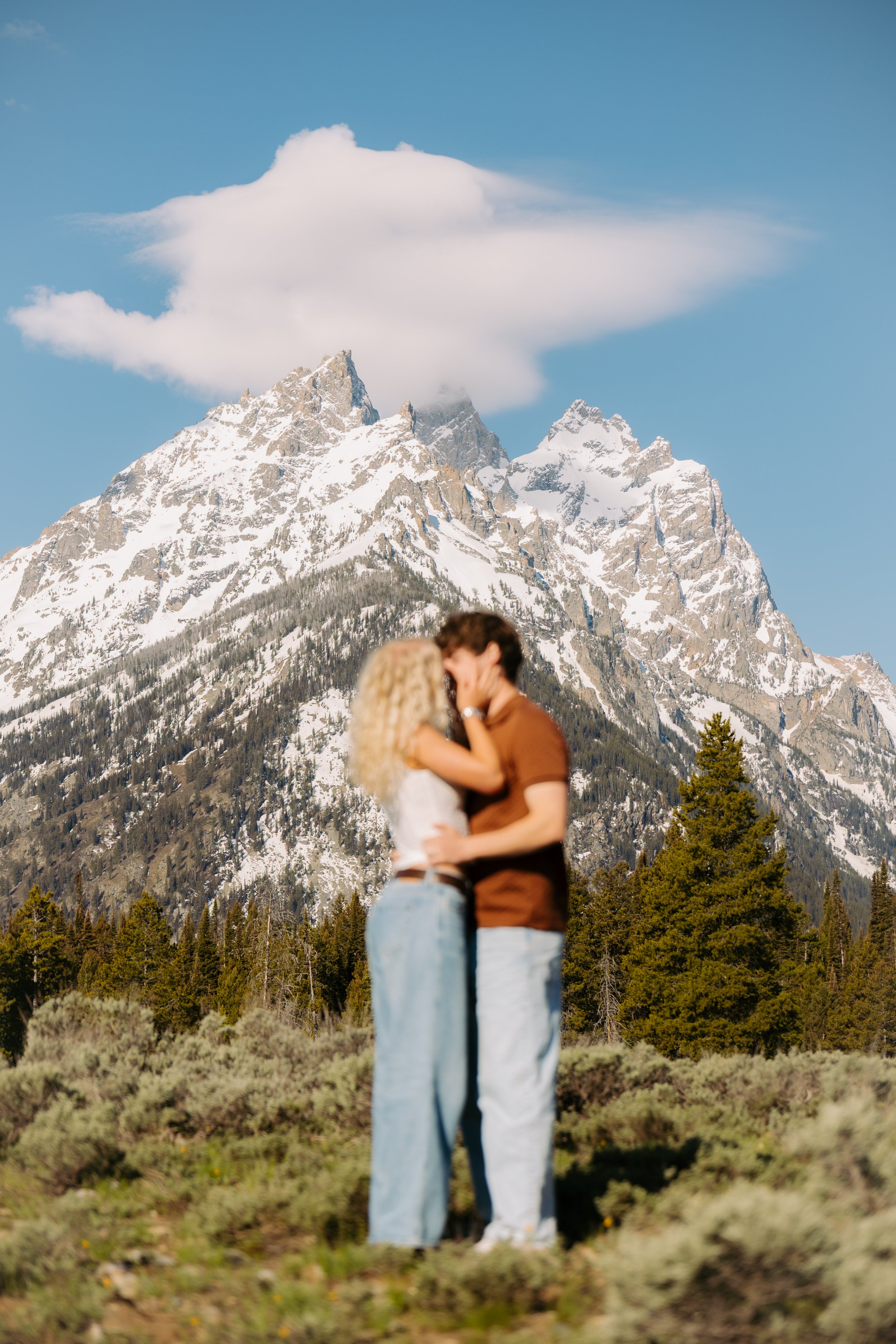 A couple kissing outdoors with snow-capped mountains and forest in the background.