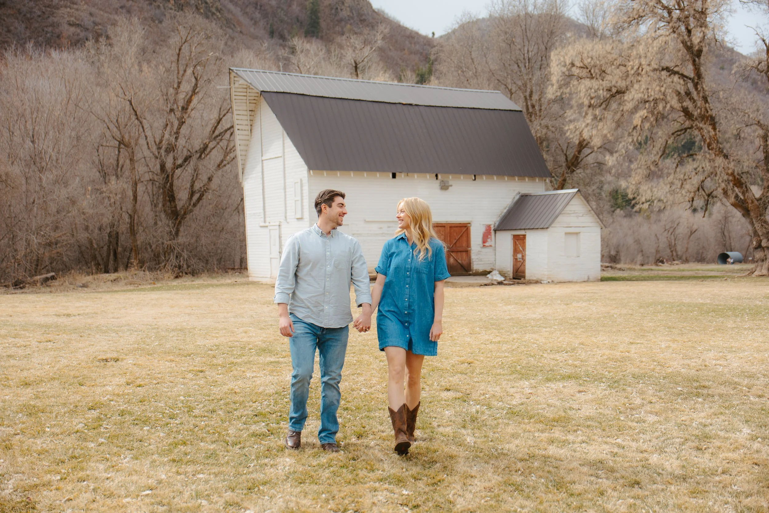 A smiling man and woman walking hand in hand on a grassy field with a white barn and leafless trees in the background, during late fall or winter.