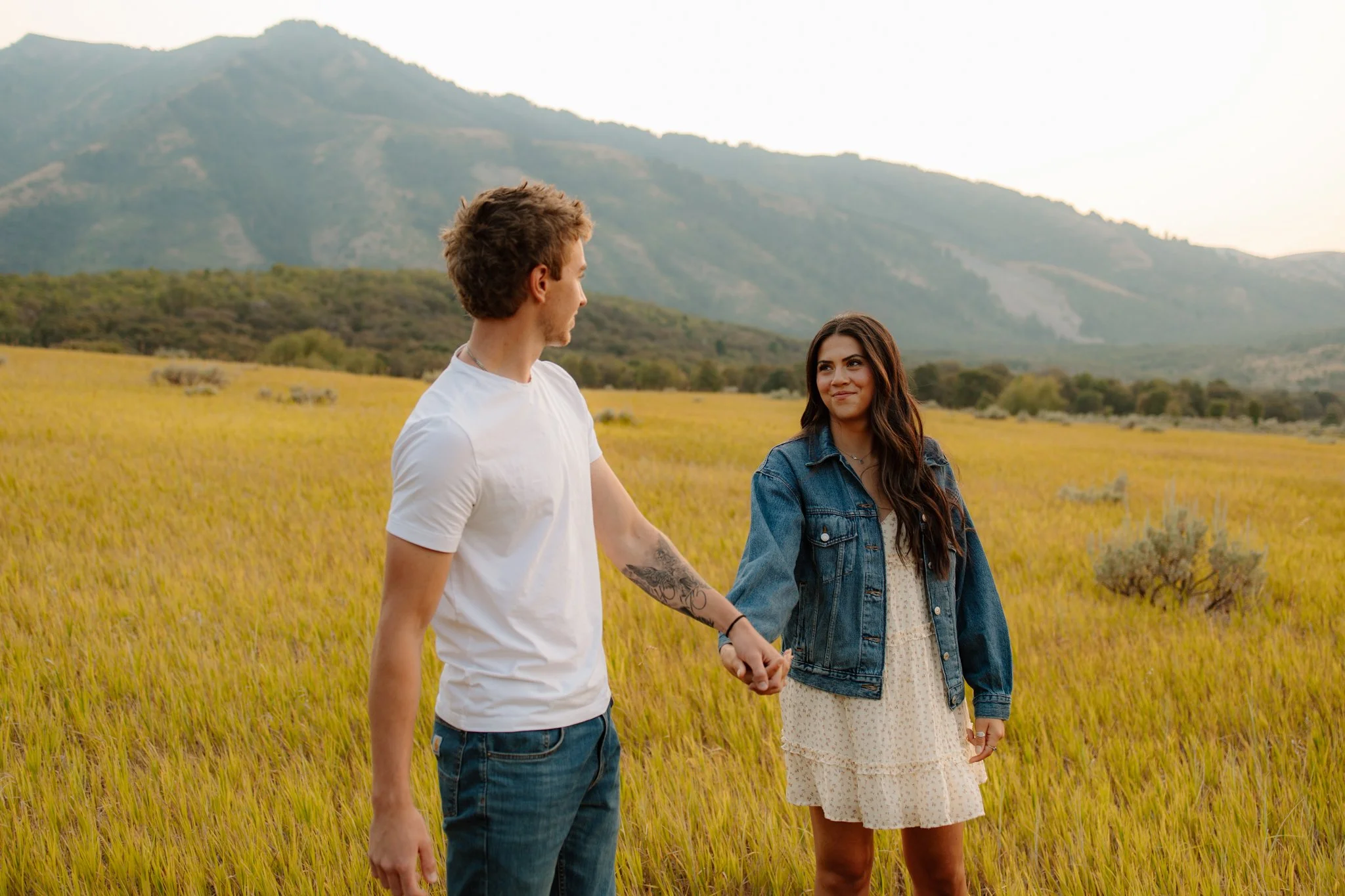 A young man and woman holding hands in a grassy field with yellow grass, mountains in the background, during sunset.