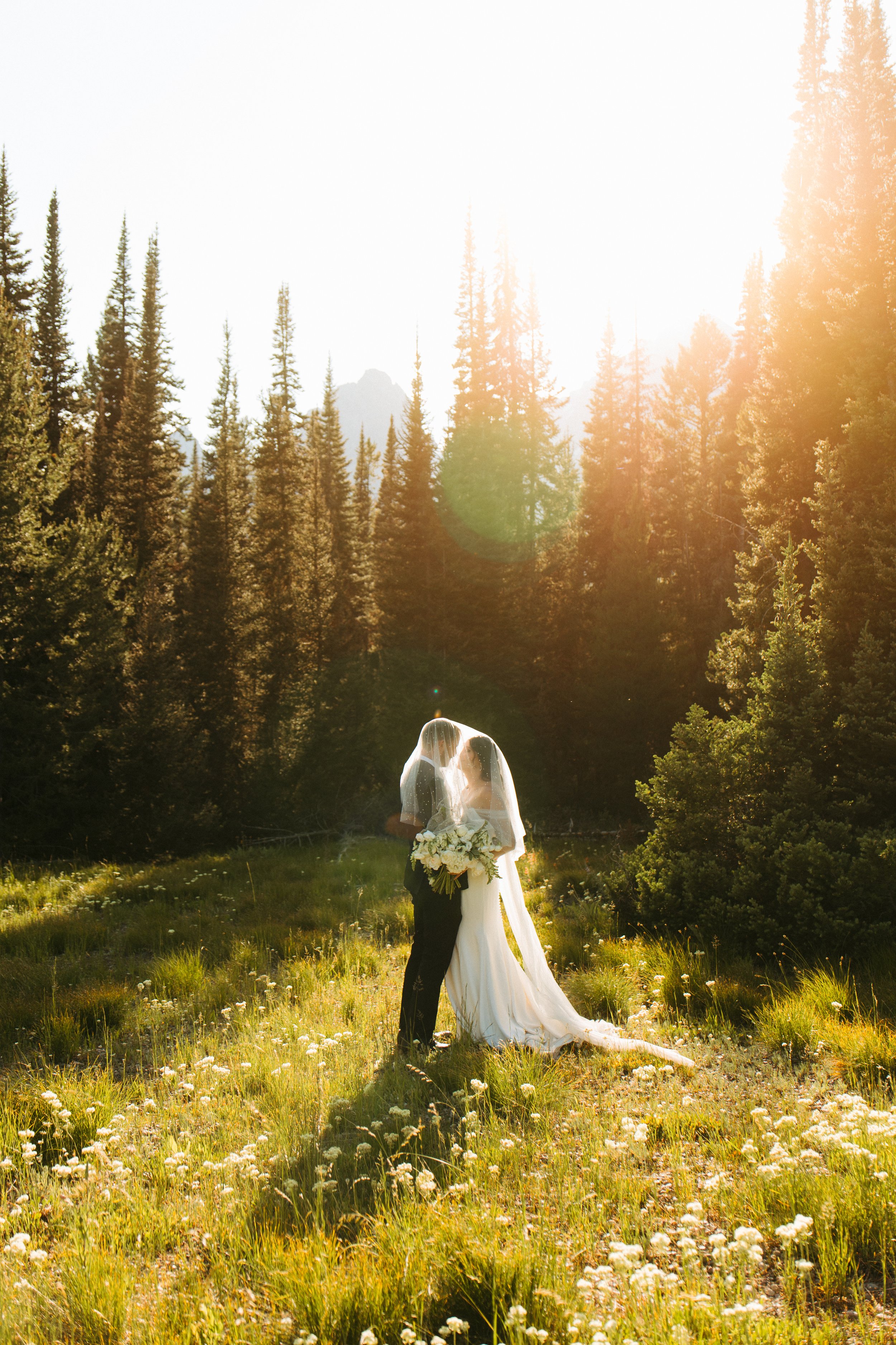A bride and groom sharing a kiss in a sunlit forest clearing with tall trees and wildflowers.