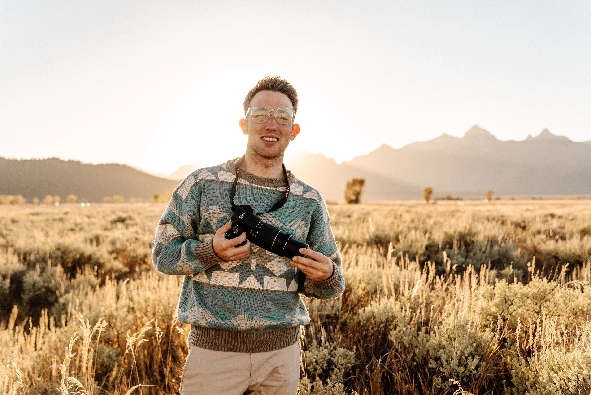 A young man holding a camera in a field at sunset with mountains in the background.