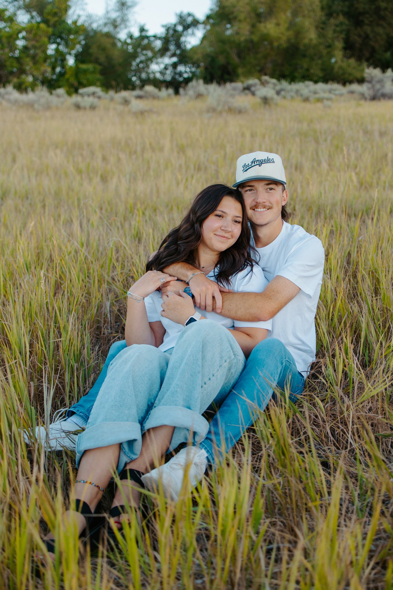 A happy young couple sitting together in a grassy field, embracing and smiling at the camera.