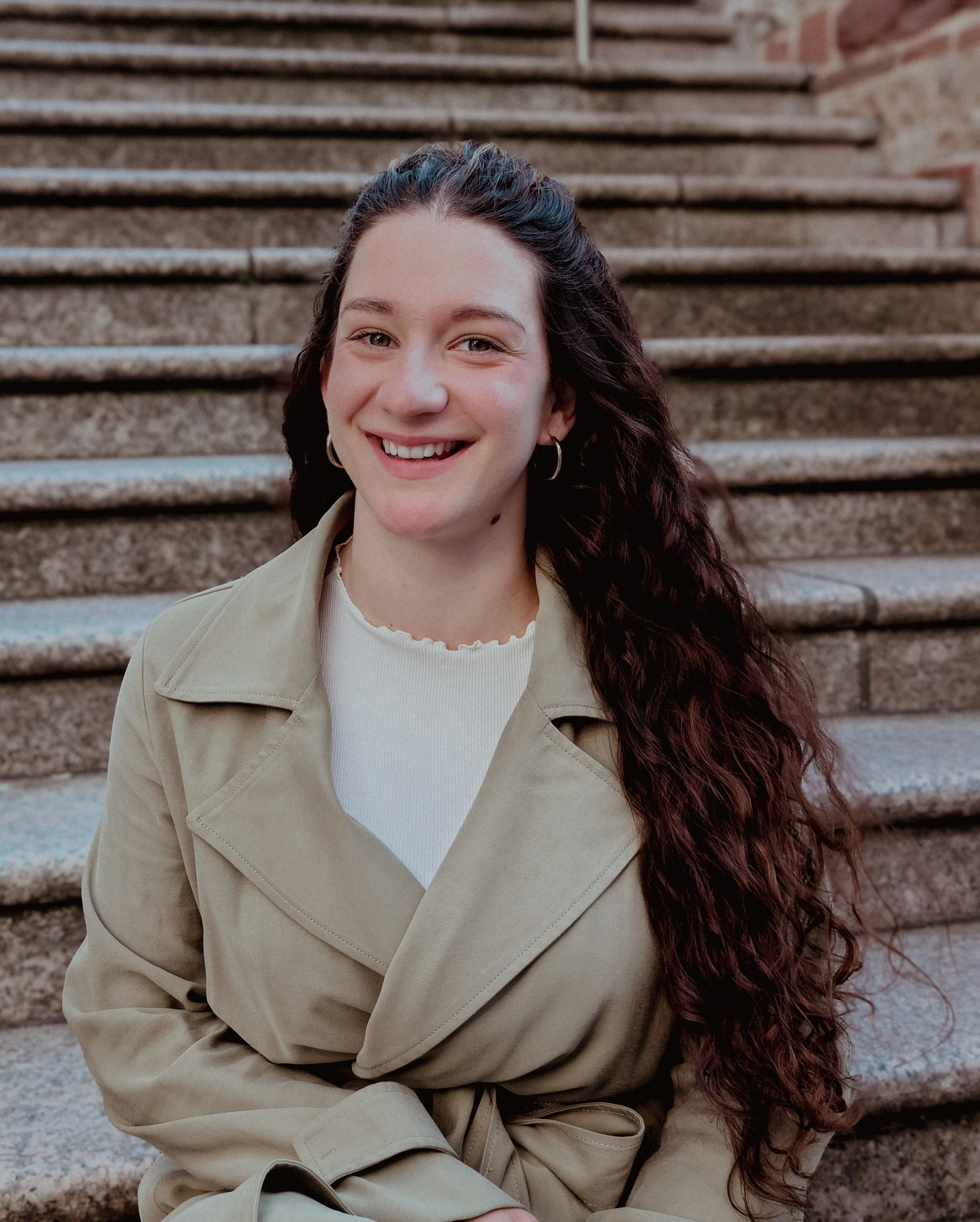 Freie Traurednerin mit dunklen, lockigen Haaren, lachend auf Steinstufen in Herrenberg sitzend, hellgrüne Jacke und weiße Oberteil, in naturalem Licht.