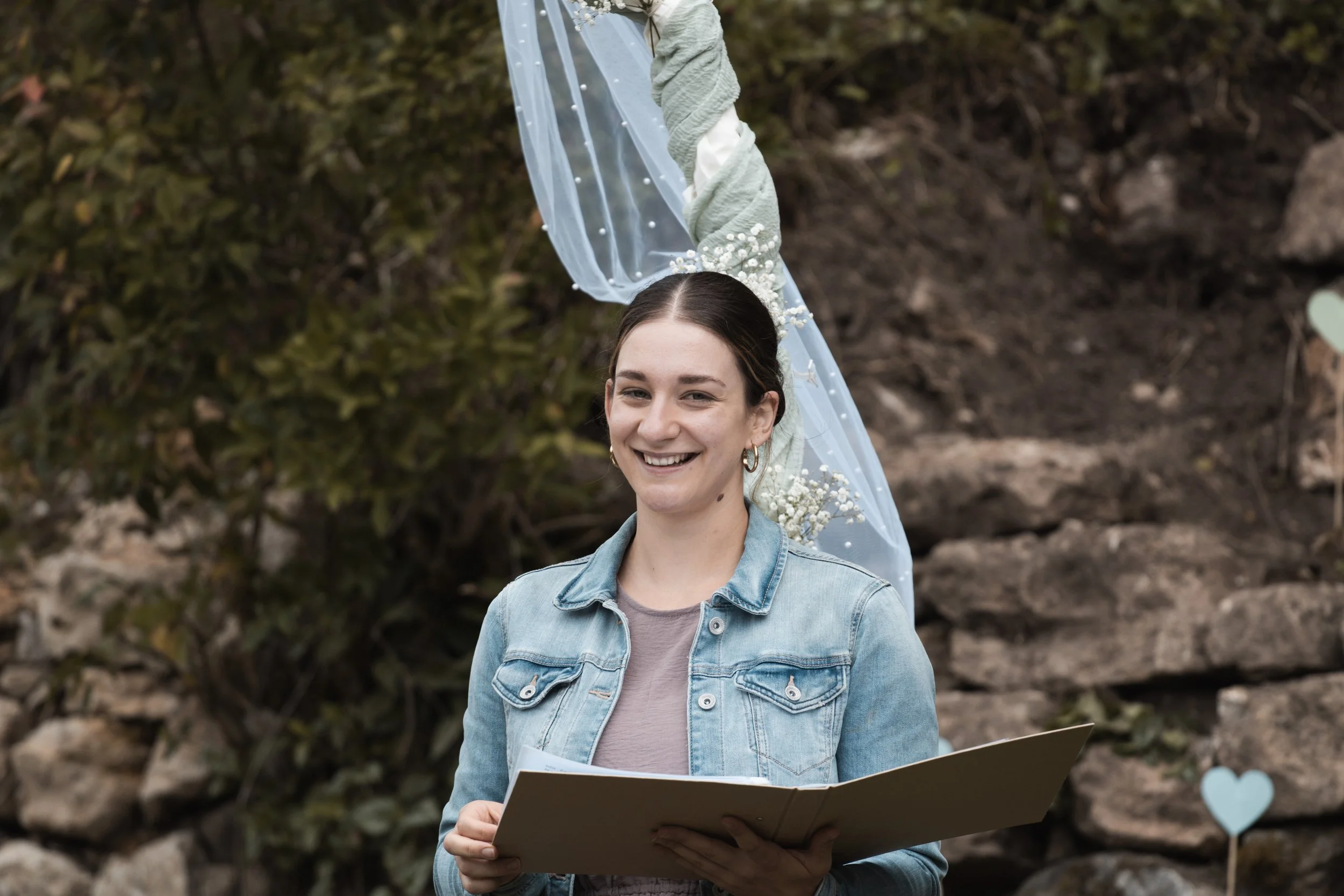 Lächelnde Traurednerin hält eine Traurede bei einer Hochzeit im Freien, im Hintergrund Steine und Bäumchen, Dekoration mit blauer Tüllstoff und kleinen weißen Blumen.