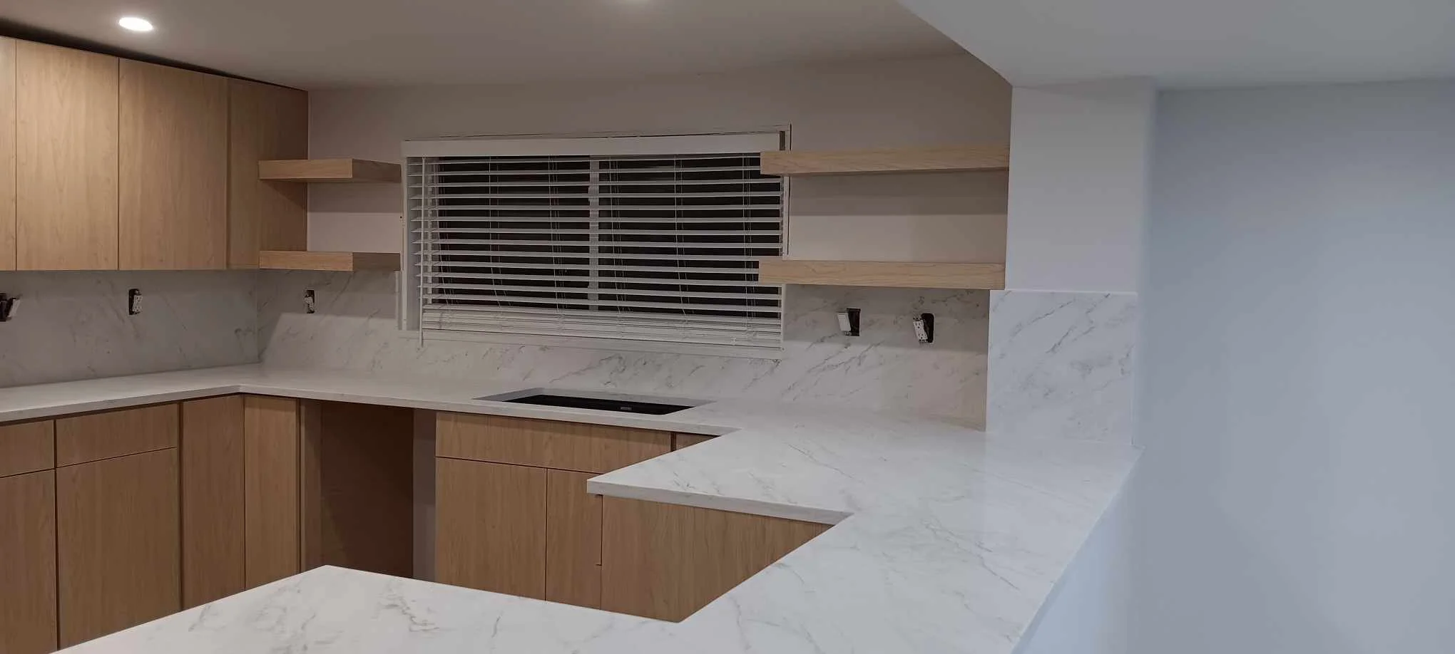 Modern kitchen with light wood cabinets, white marble countertops, and a window with blinds.