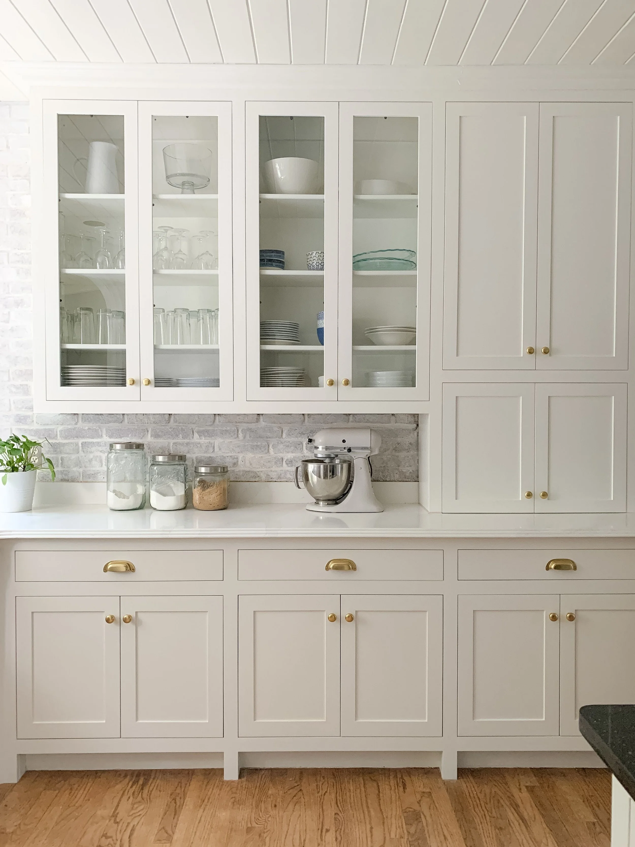 A white kitchen cabinet with glass doors and brass handles, containing glasses, plates, and bowls, with countertop items including jars, a stand mixer, and a potted plant, against a brick wall and wooden floor.