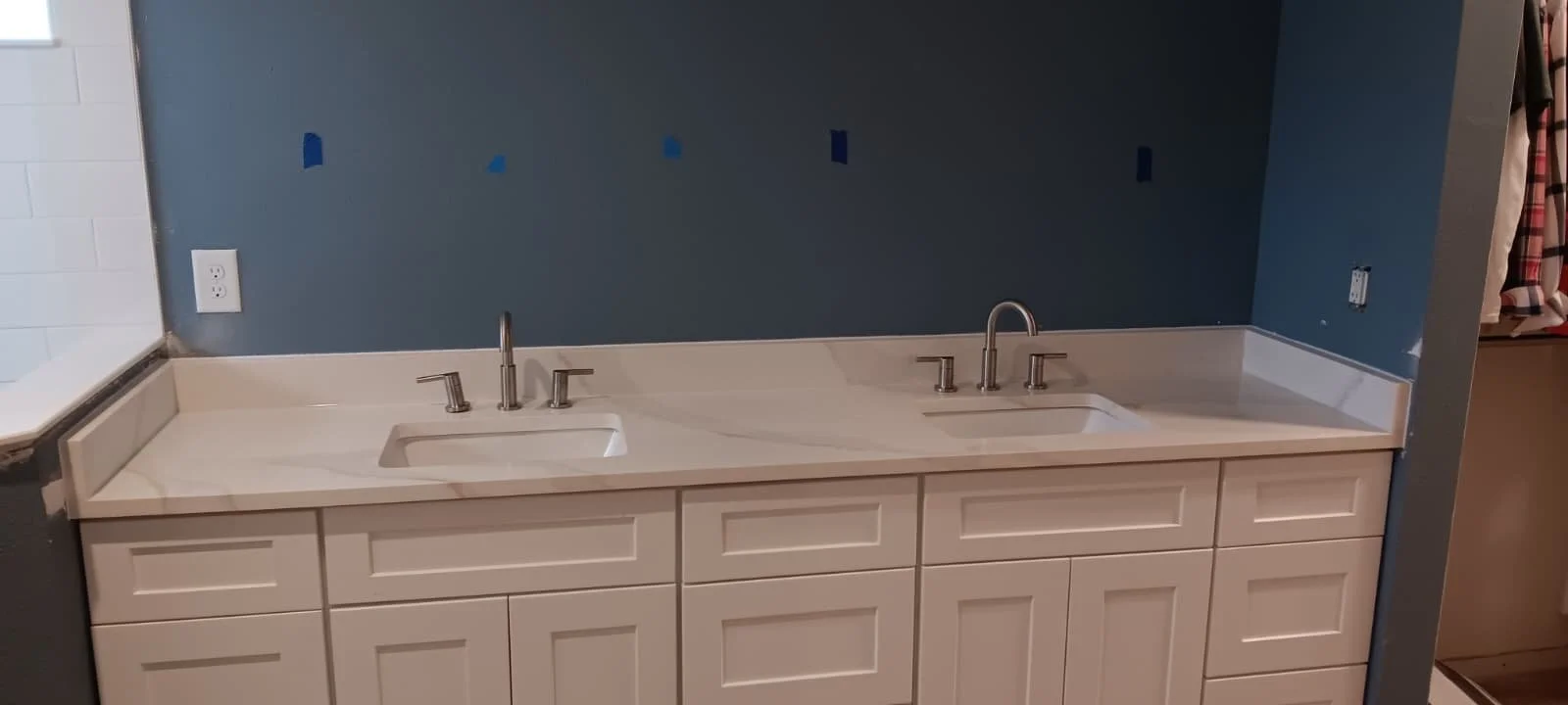 Kitchen sink cabinet with two white sinks and stainless steel faucets against a blue wall with some blue painter's tape.